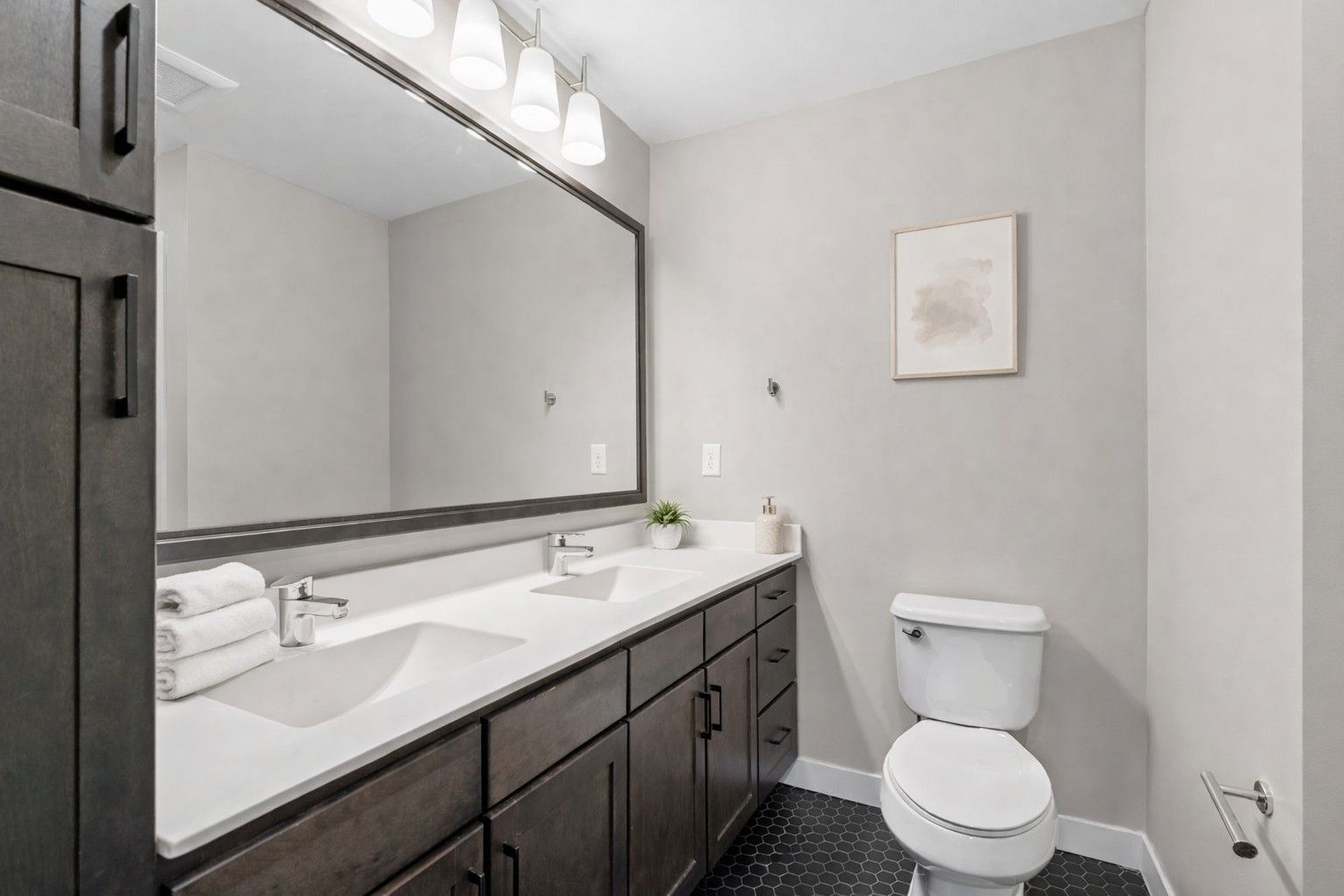 A modern bathroom here at Maverick featuring a double vanity with dark cabinets, white countertops, a large mirror, and a white toilet.