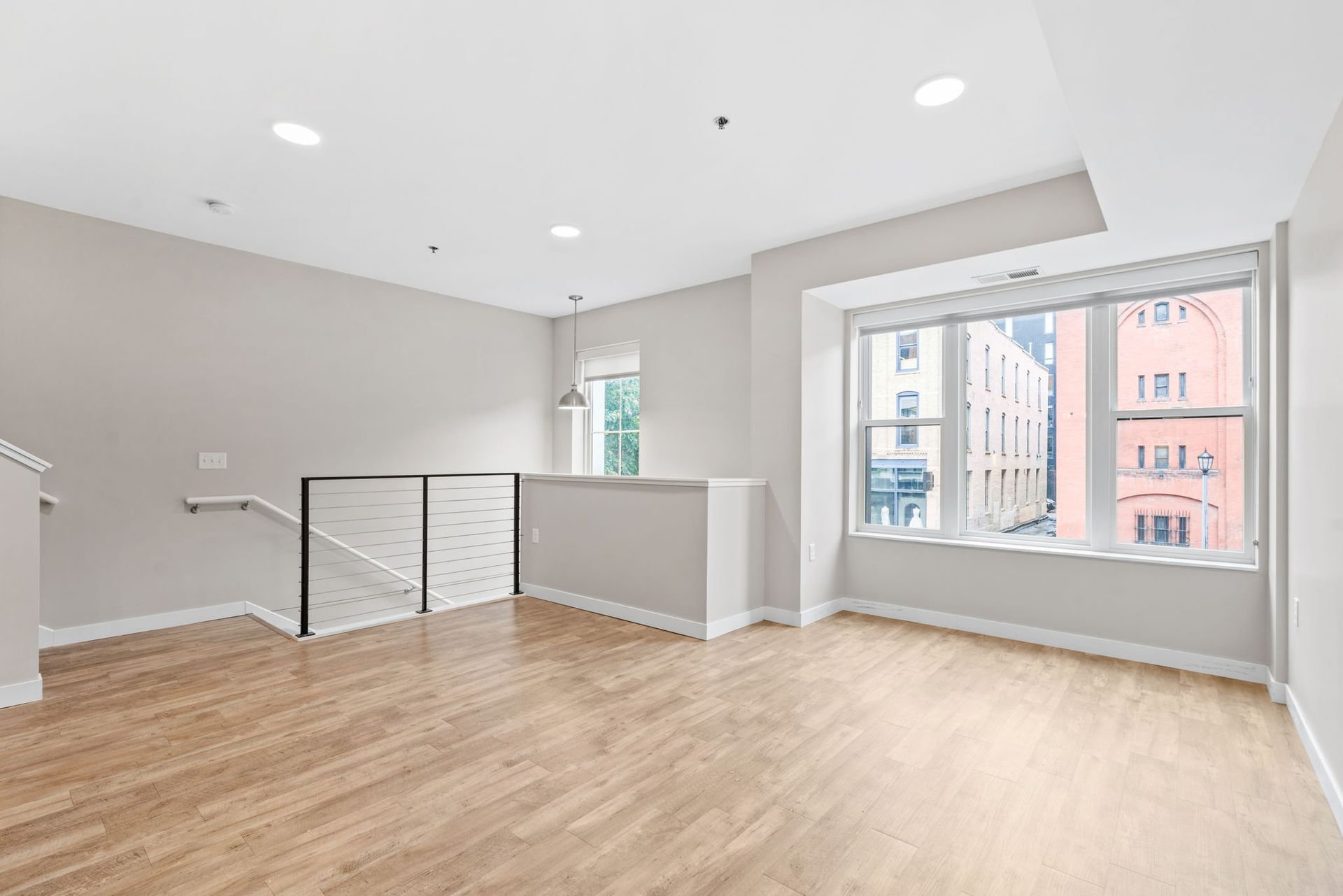 A bright, empty room here at Maverick with light wood floors, beige walls, a white staircase railing, and large windows facing a city view.