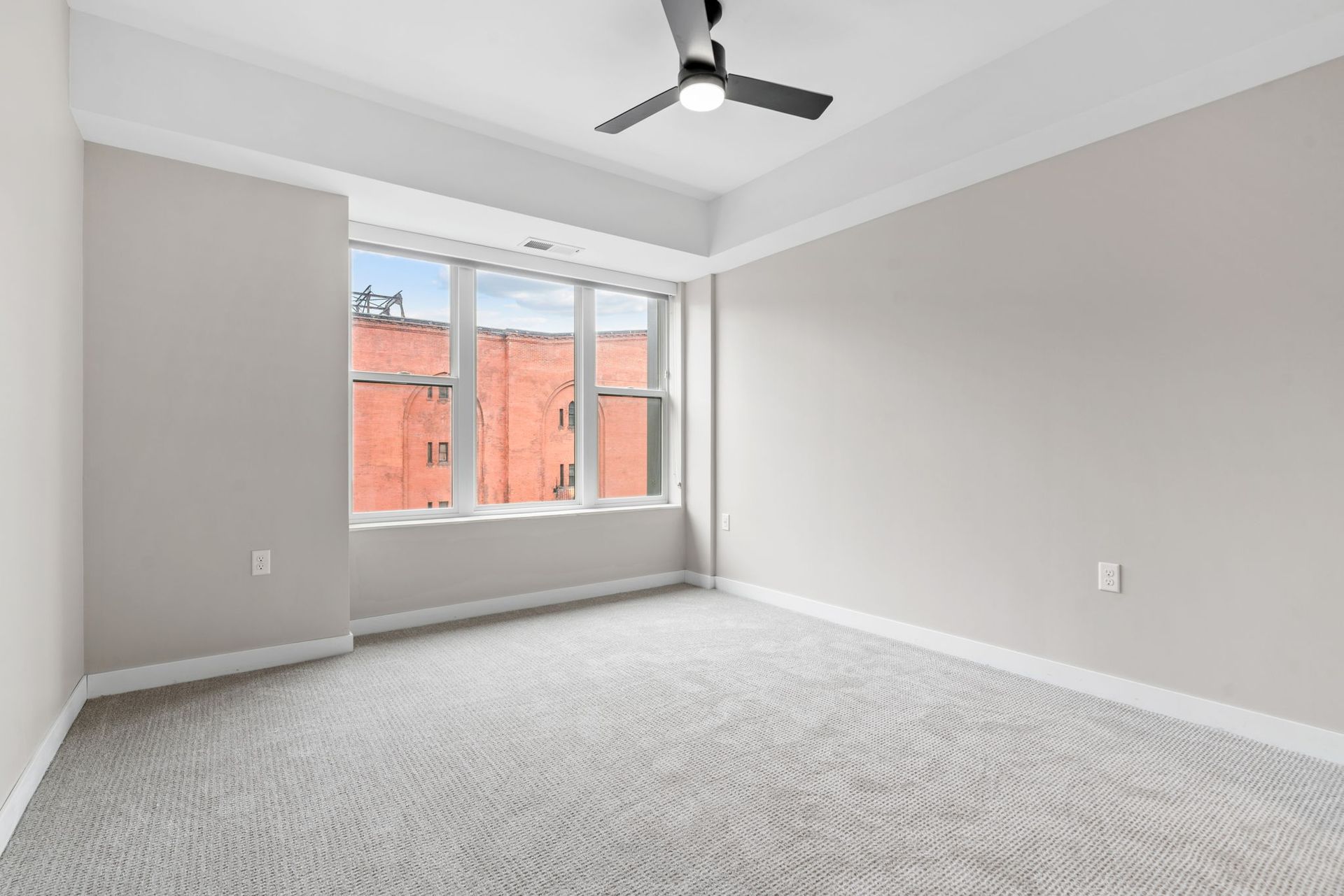 An empty bedroom here at Maverick with light gray walls, white trim, gray patterned carpet, a ceiling fan, and a large window.