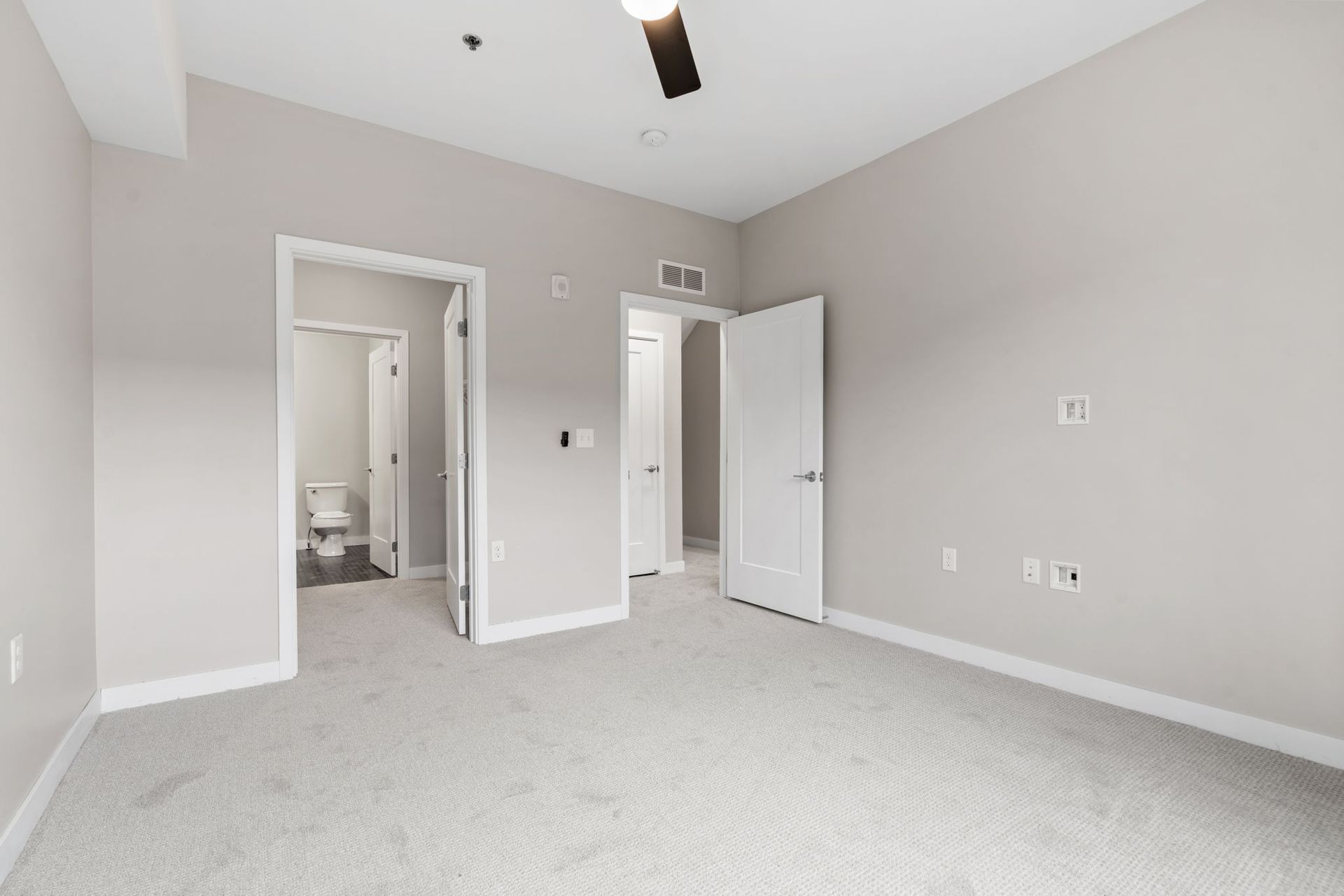 An empty room with light gray carpet and walls, featuring two open doorways revealing a bathroom and a closet.