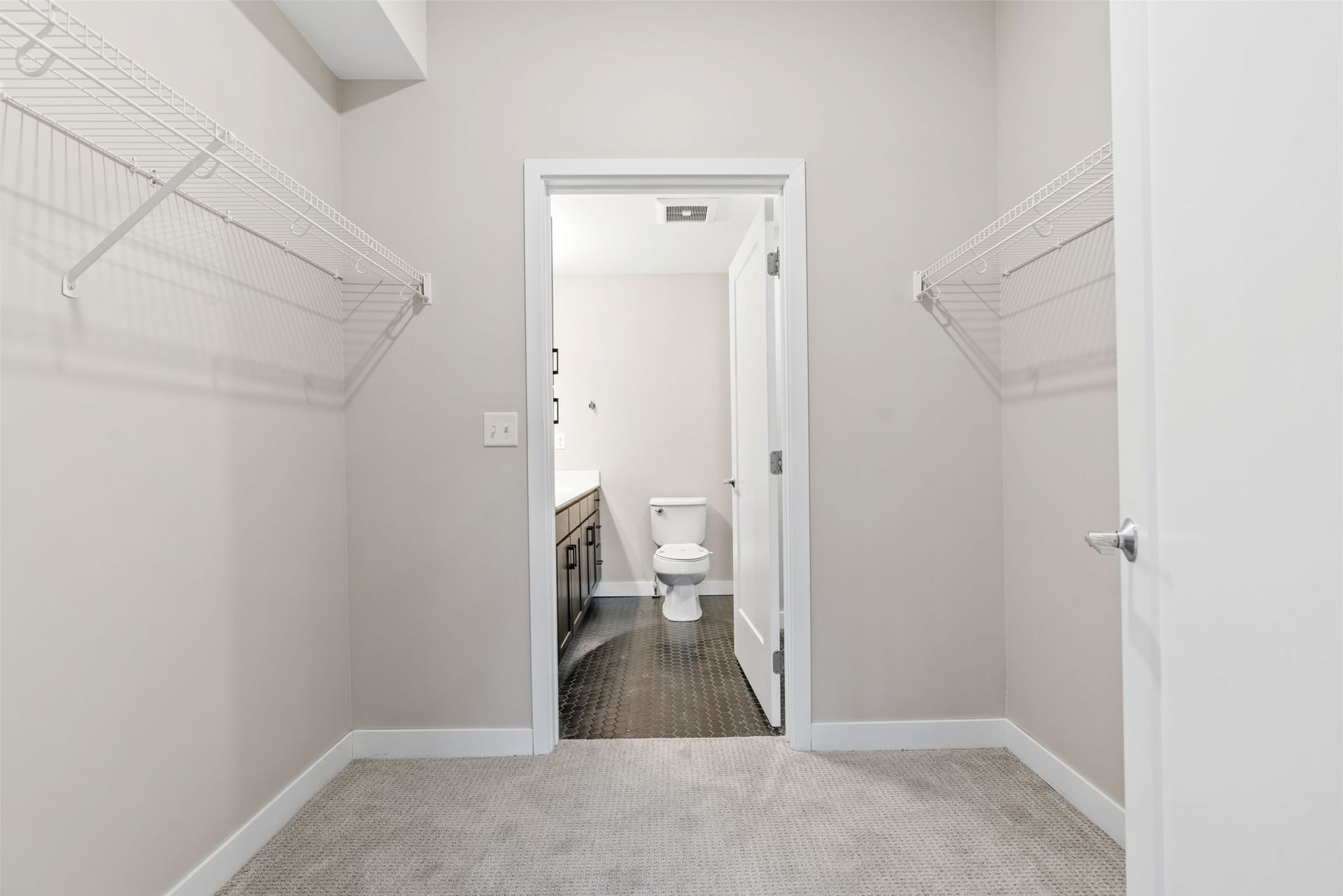 A walk-in closet with wire shelving on both sides, leading to a doorway that reveals a small bathroom with a toilet.