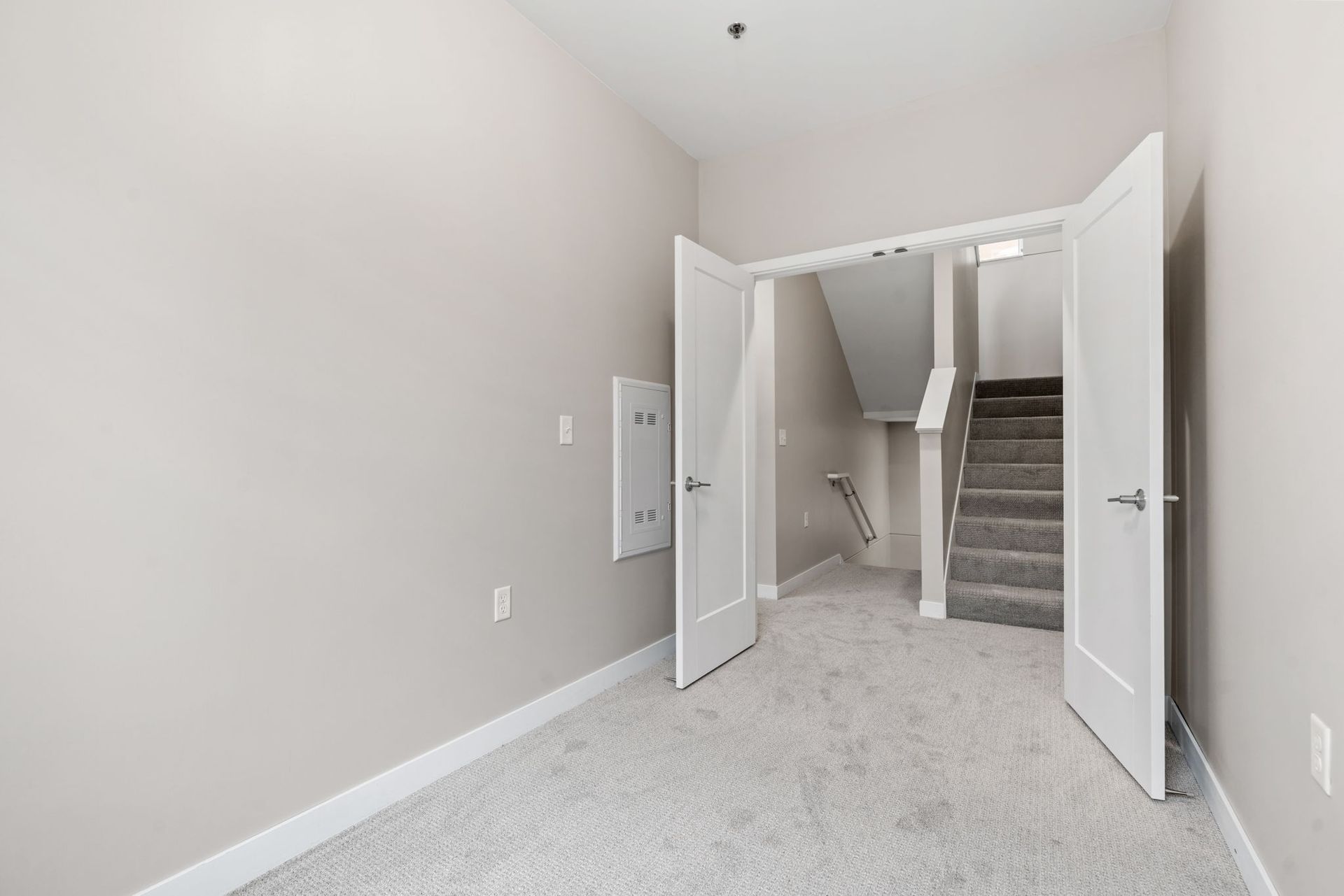 An interior view of a room with light grey carpet and walls, showing an open double door leading to a carpeted staircase.