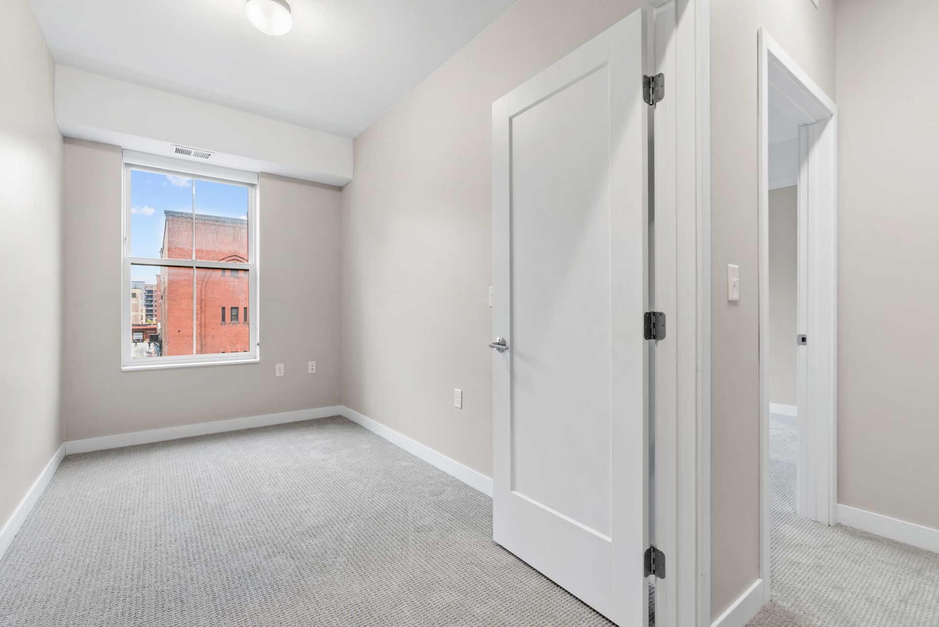 An empty room with gray carpet, neutral walls, a window overlooking a brick building, and a white interior door.