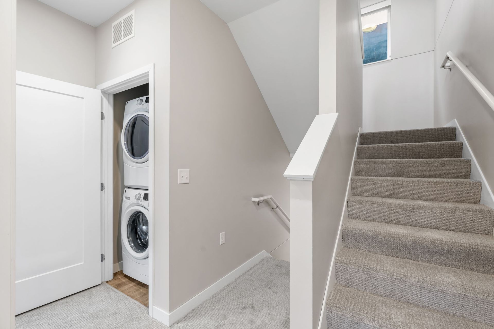 A beige interior view showing a white door, a stacked washer and dryer unit, and a carpeted staircase with a handrail.