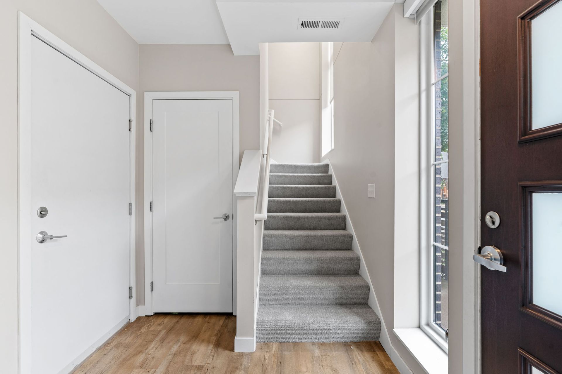 A brightly lit entryway with light wood flooring, two white doors, and a carpeted staircase leading to an upper level.