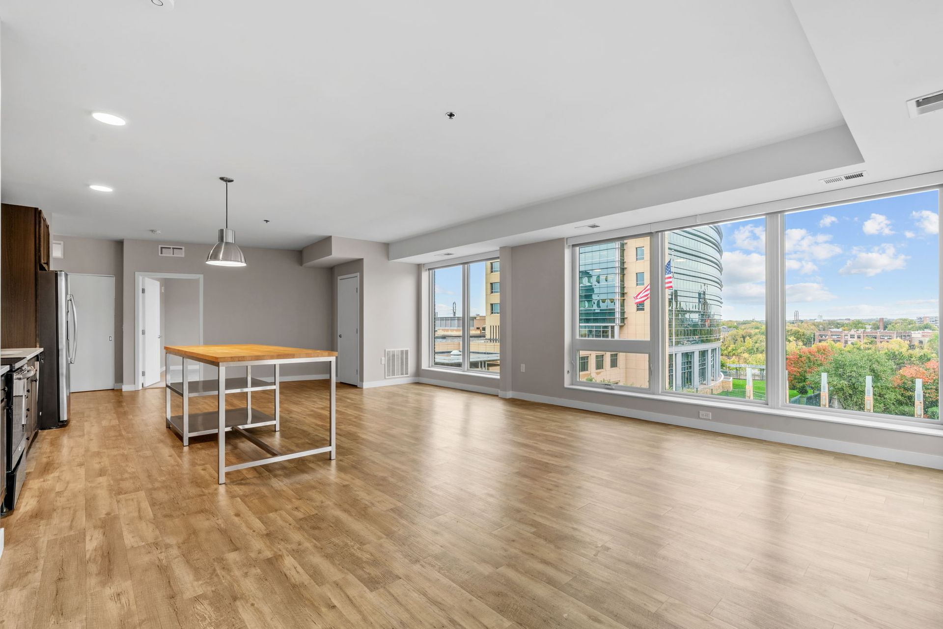 A wide, sunlit apartment living area with light wood floors, a kitchen island, and large windows with a city view.