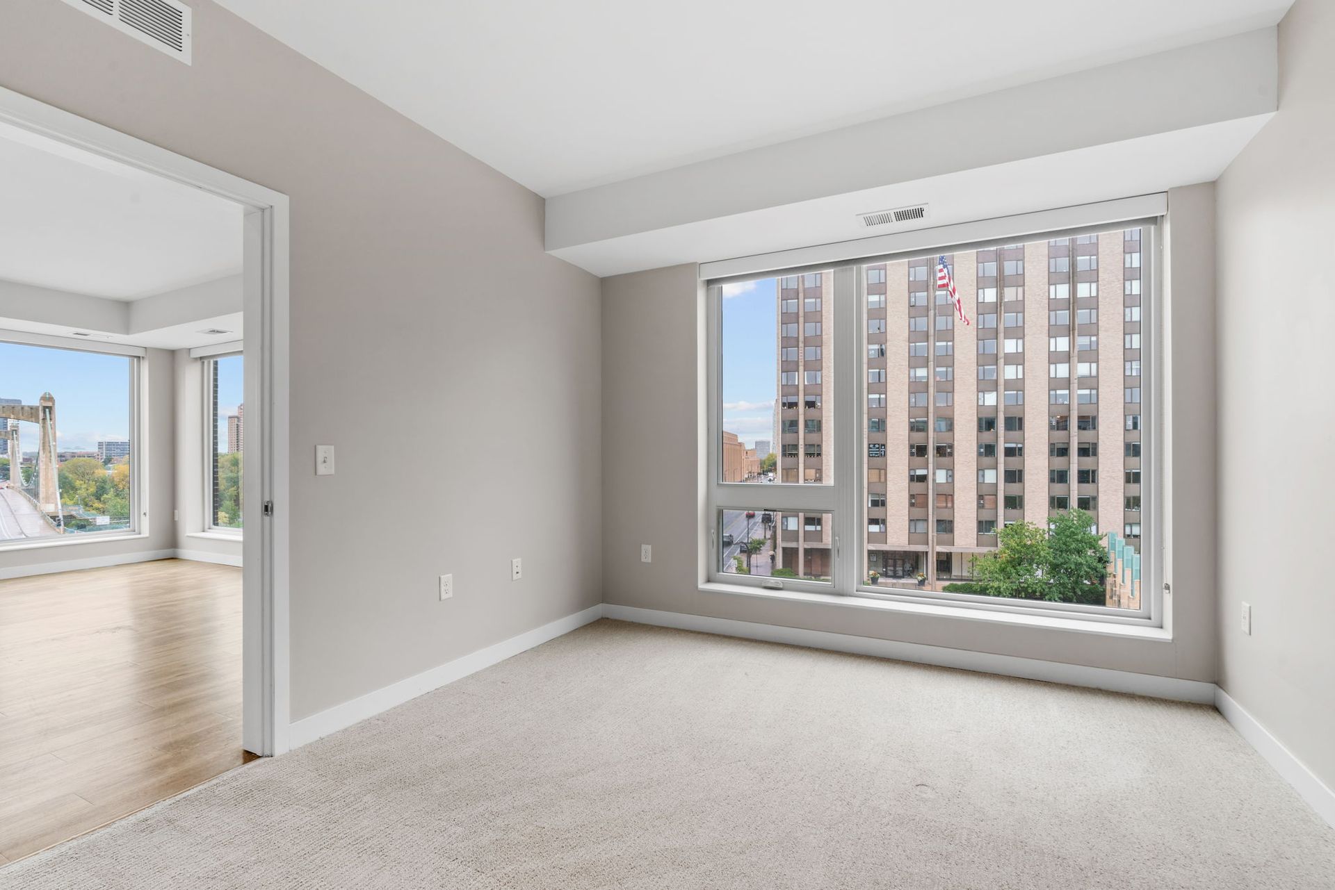 Empty room with beige carpet, light gray walls, a doorway to another room, and a large window facing city buildings.