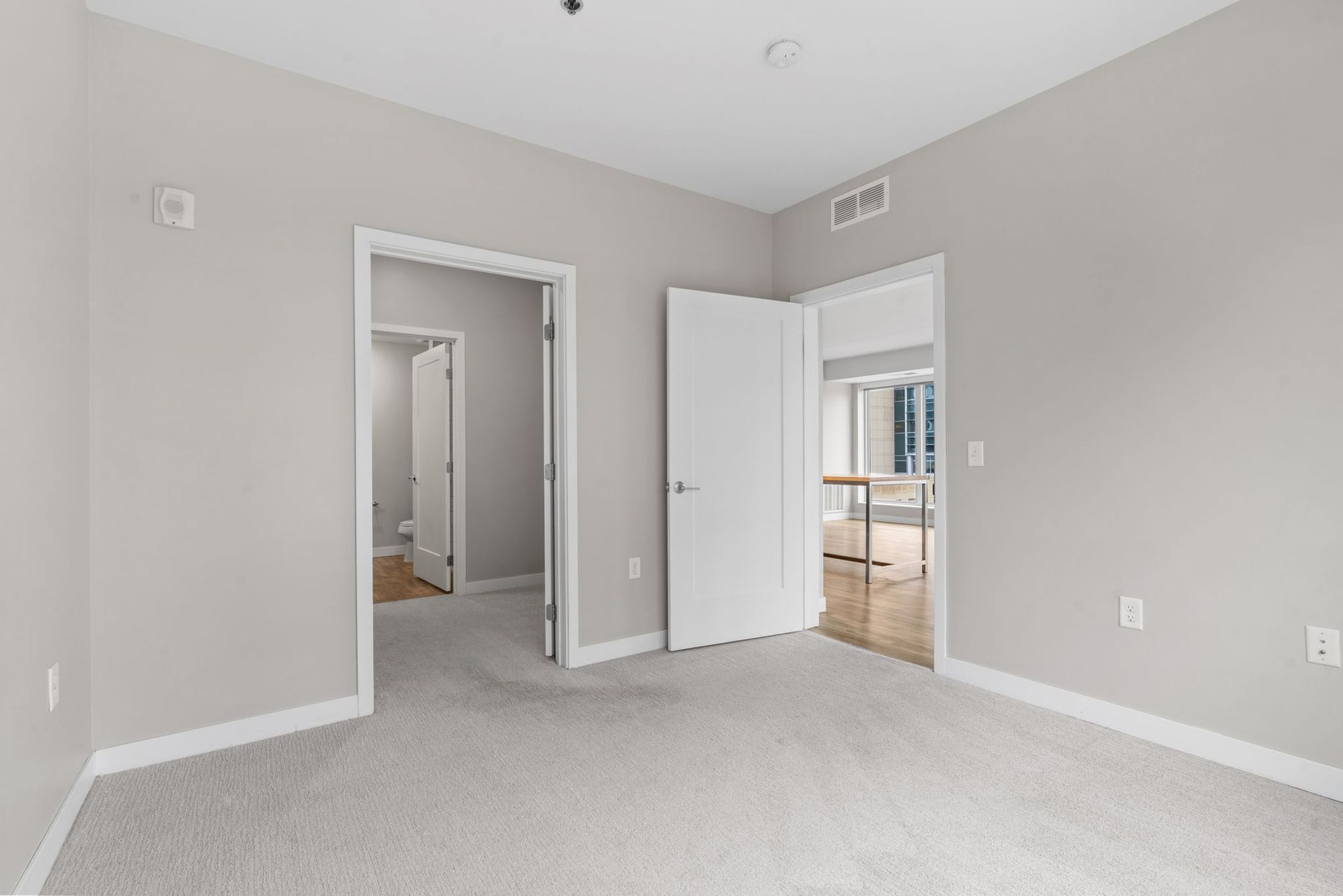 Empty bedroom with gray carpet, light gray walls, white trim, and two open doorways leading to other rooms.