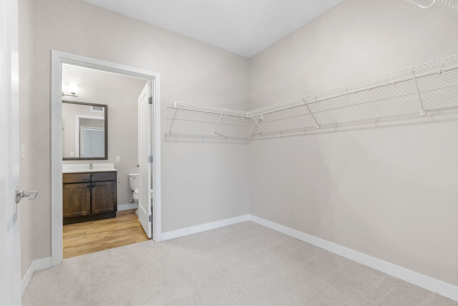 An empty walk-in closet with beige carpet, white walls, and wire shelving, looking into a bathroom with a wood vanity.