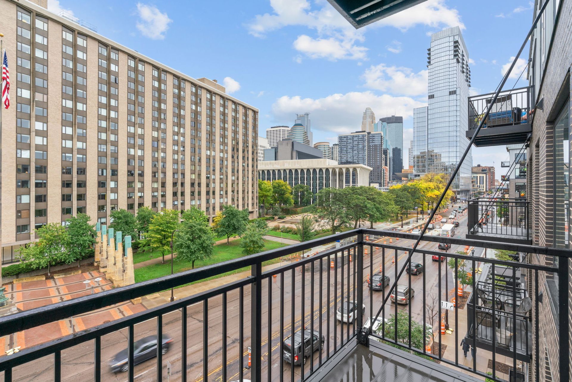 A view of a city skyline and tree-lined street from a balcony with a black metal railing under a blue sky.