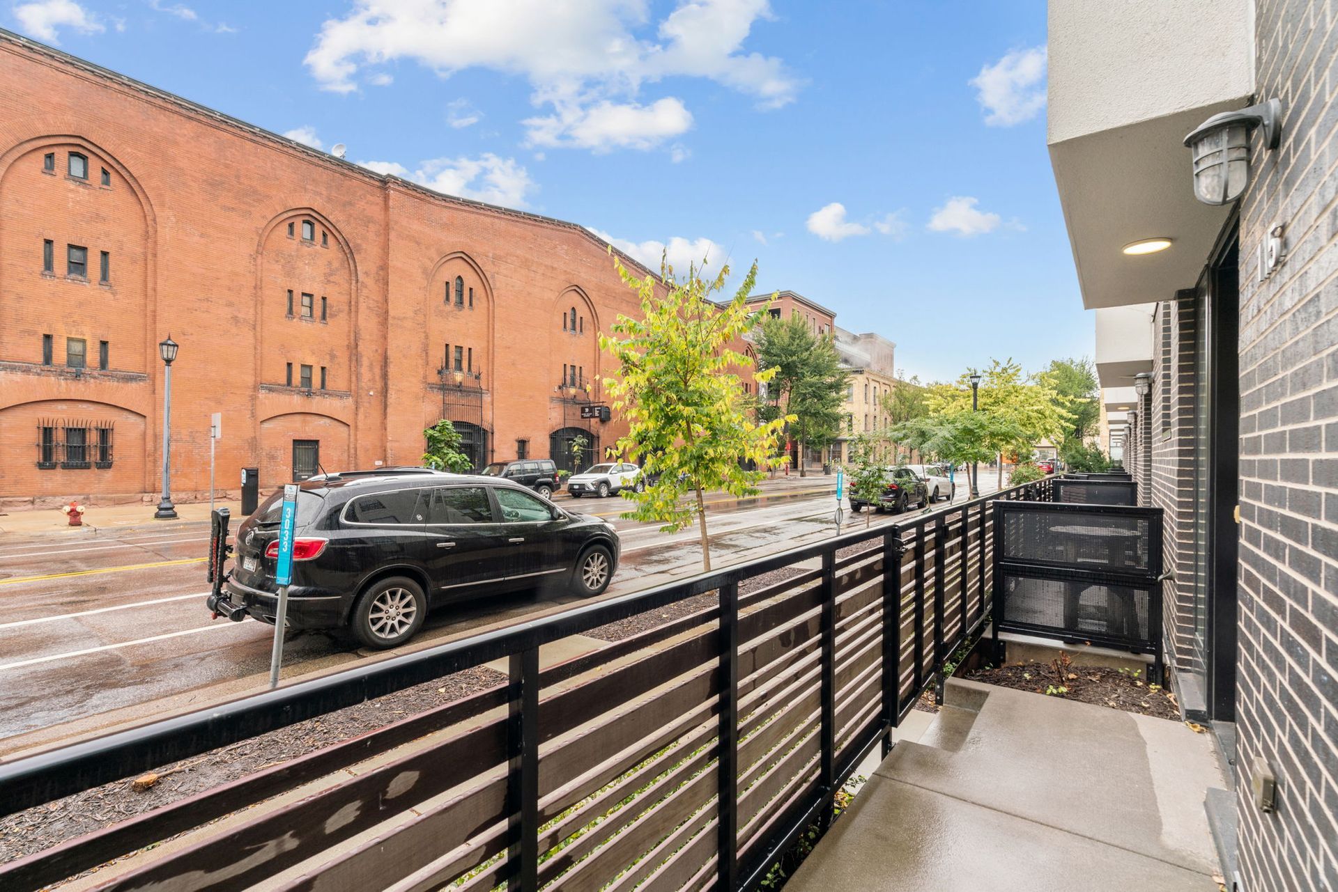 An outdoor balcony view featuring a black metal railing, overlooking a street with a black SUV and a large brick building.