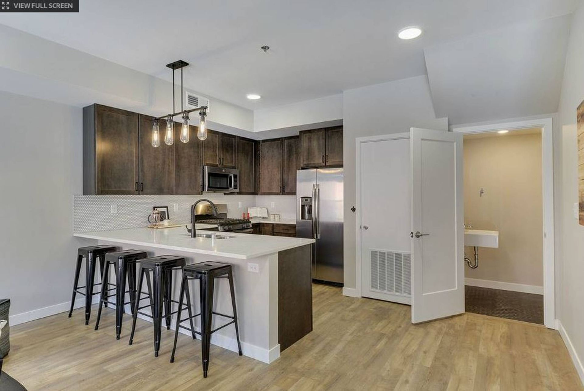 A modern kitchen here at Maverick with dark wooden cabinets, a white island with four black stools, and an open doorway to a bathroom.