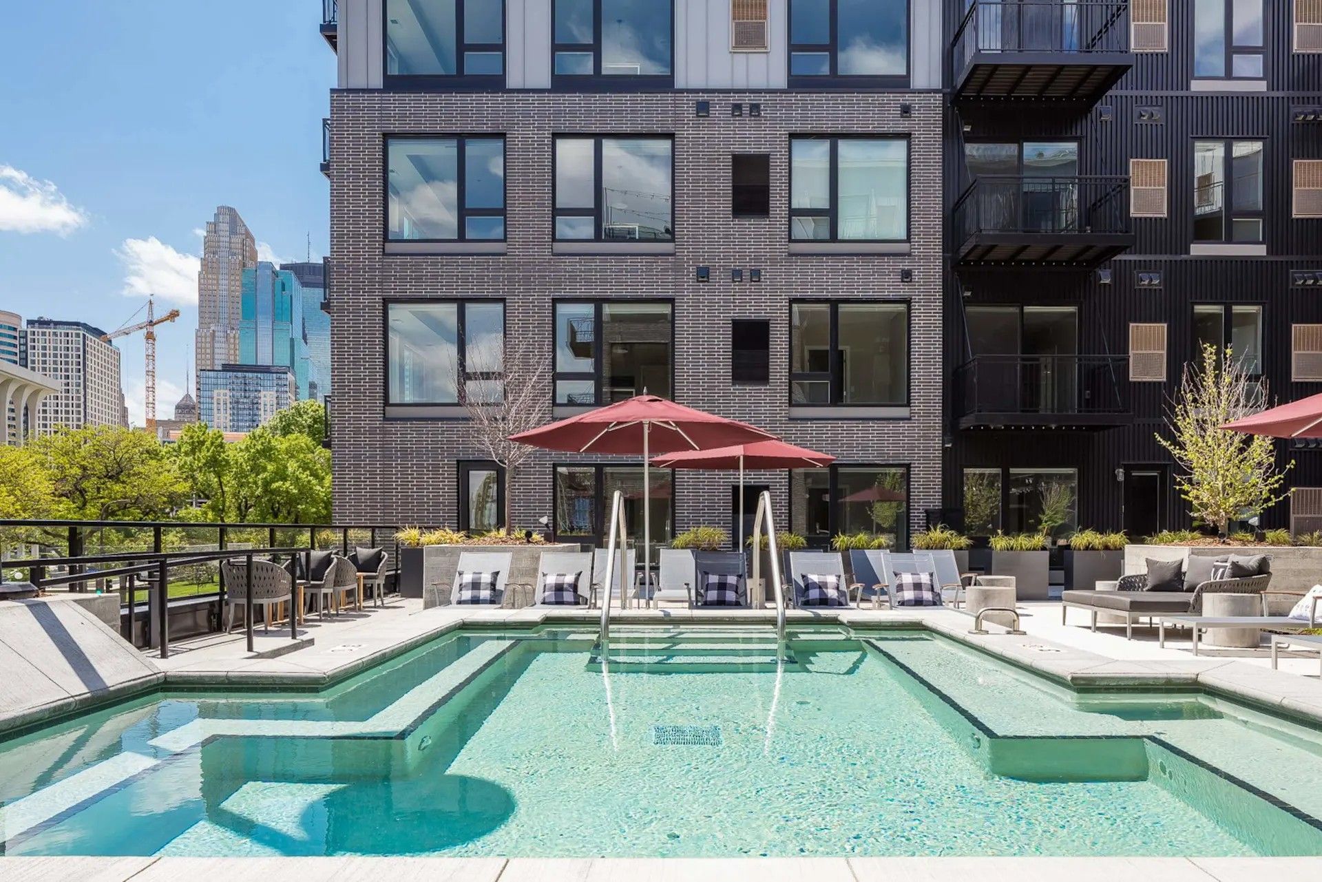 A rooftop swimming pool area with lounge chairs, umbrellas, and a brick apartment building in an urban setting.