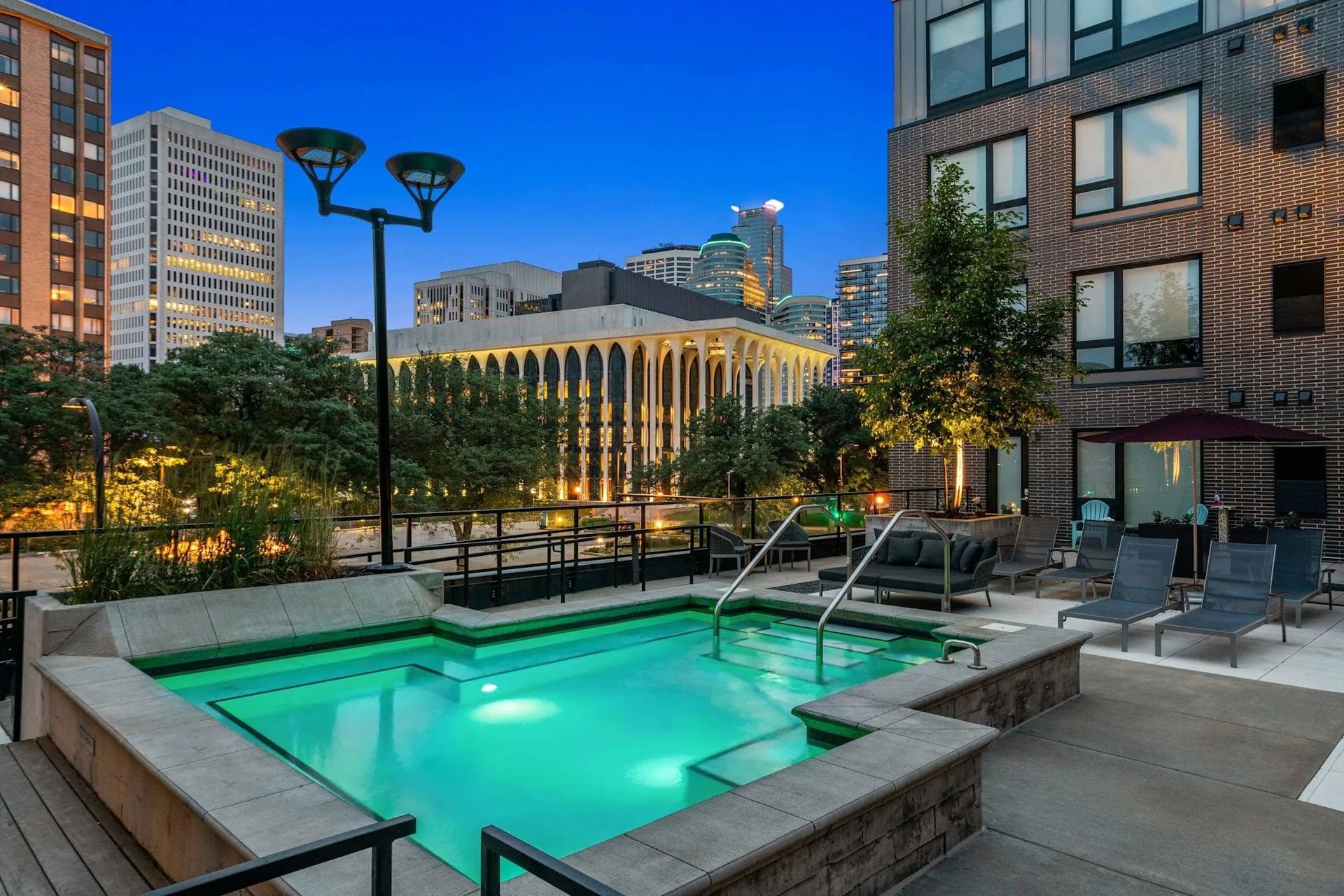 An illuminated outdoor hot tub on a building rooftop deck at dusk, surrounded by trees and city skyline views.