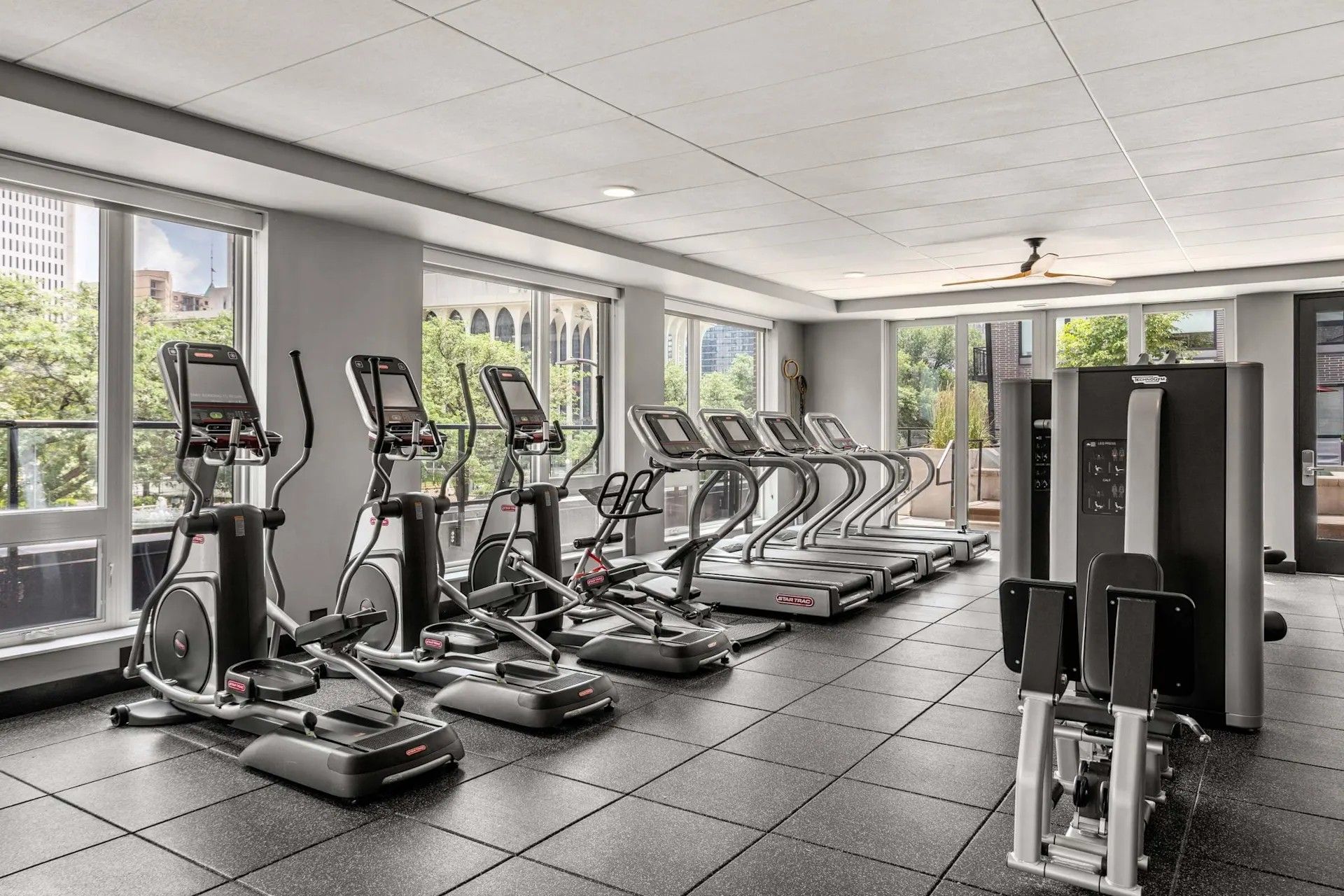 A bright fitness center featuring a row of elliptical machines and treadmills in front of large windows.