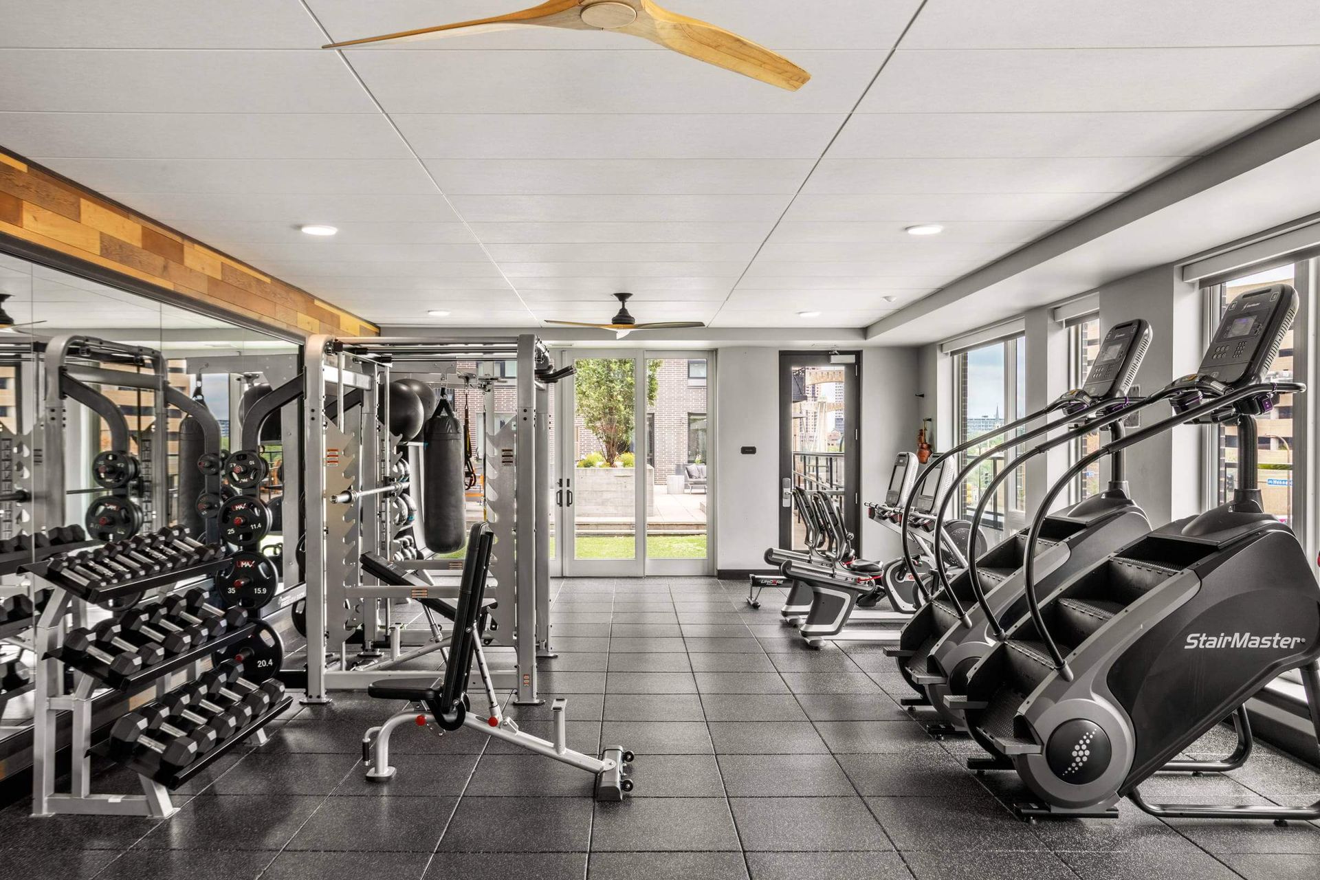 Modern indoor gym featuring dumbbell racks, strength training equipment, and stair climbers on a dark rubber floor.