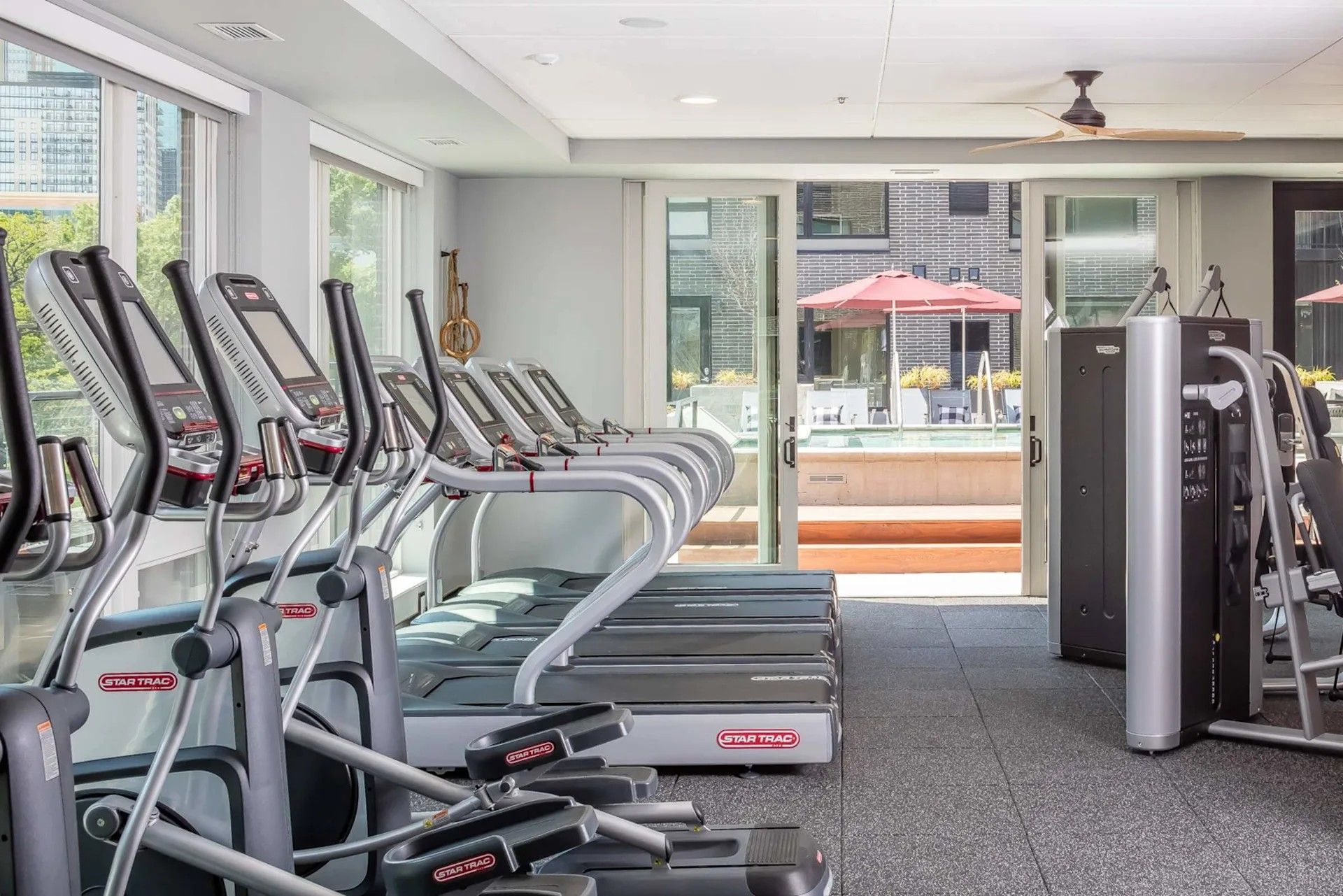 A brightly lit fitness center featuring rows of treadmills and a strength training machine with an outdoor patio view.