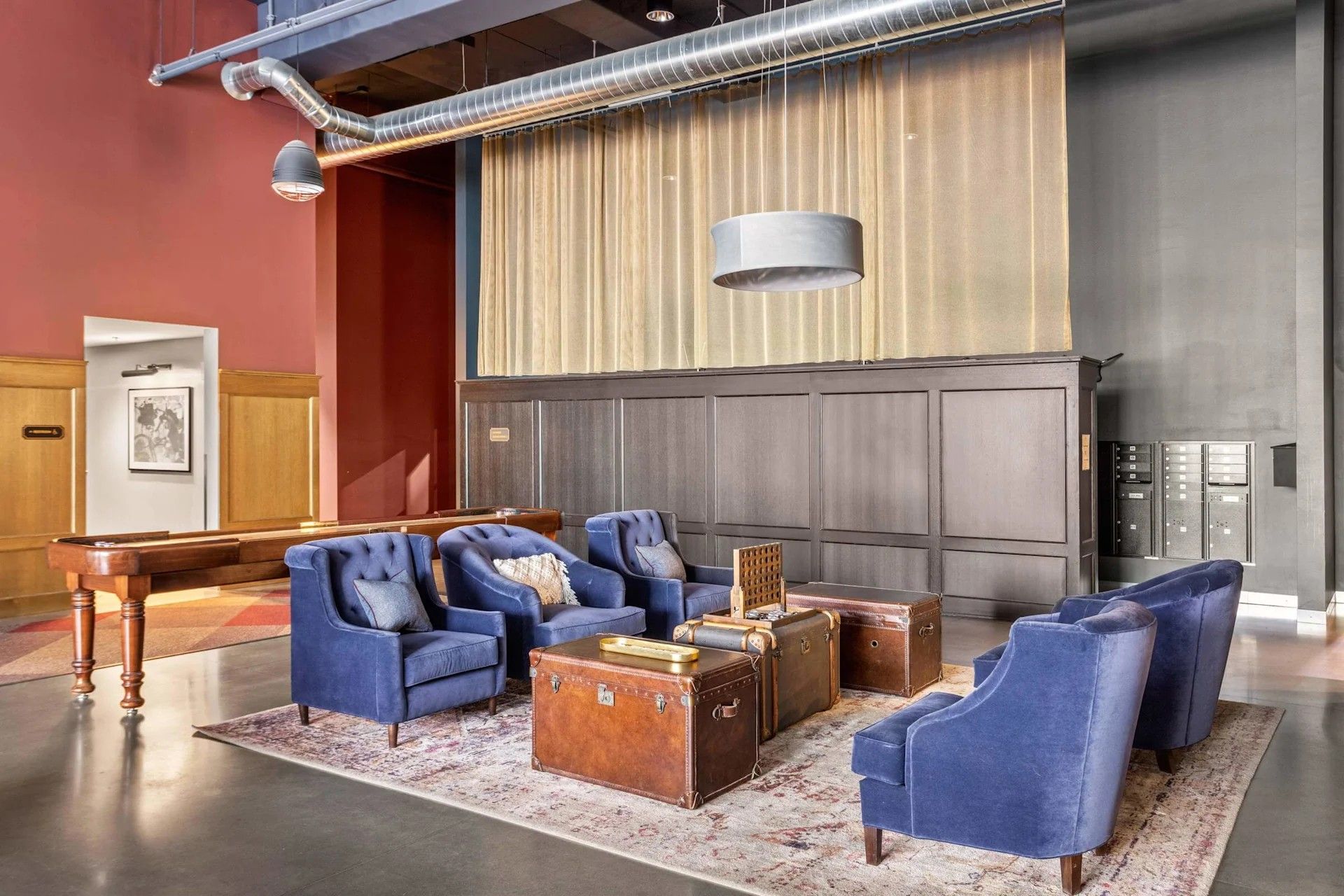 A lobby with blue armchairs, brown leather trunks as tables on a rug, a shuffleboard table, and a metallic curtain wall.