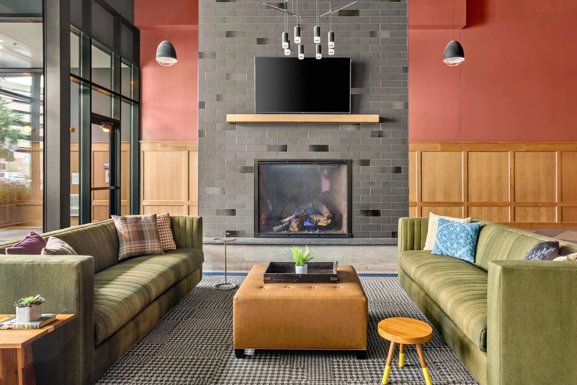 Modern lounge area featuring two olive green sofas, a central brown ottoman, and a brick fireplace with a mounted TV.