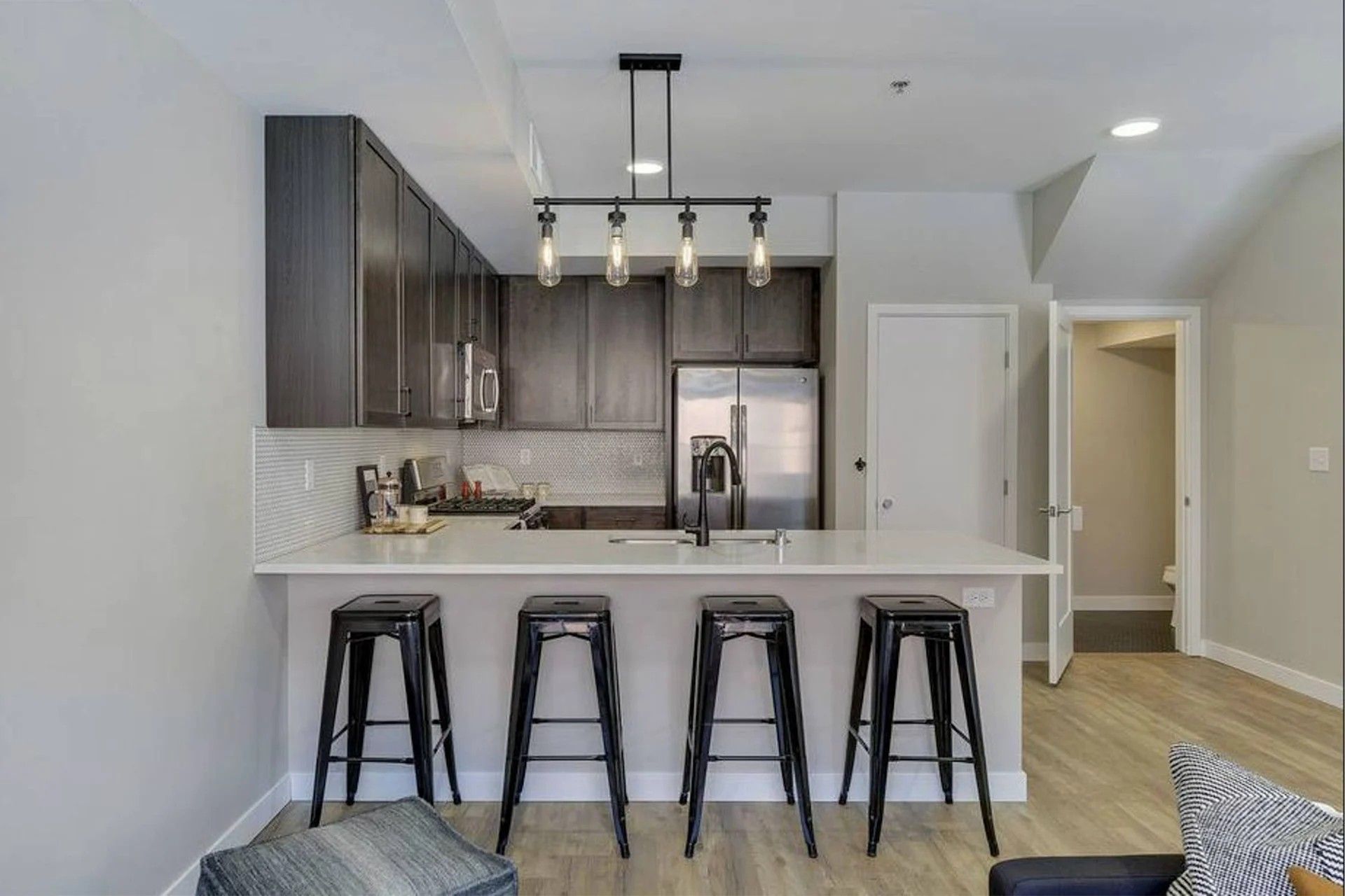 A modern kitchen here at Maverick features dark cabinetry, a white countertop with four black bar stools, and a linear pendant light.