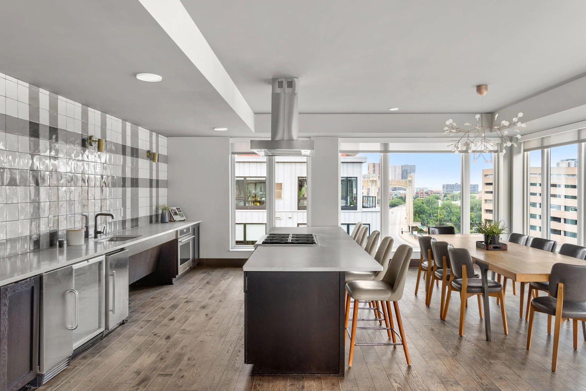 A modern kitchen featuring a dark island with bar stools, a long table, stainless appliances, and a plaid-tiled wall.