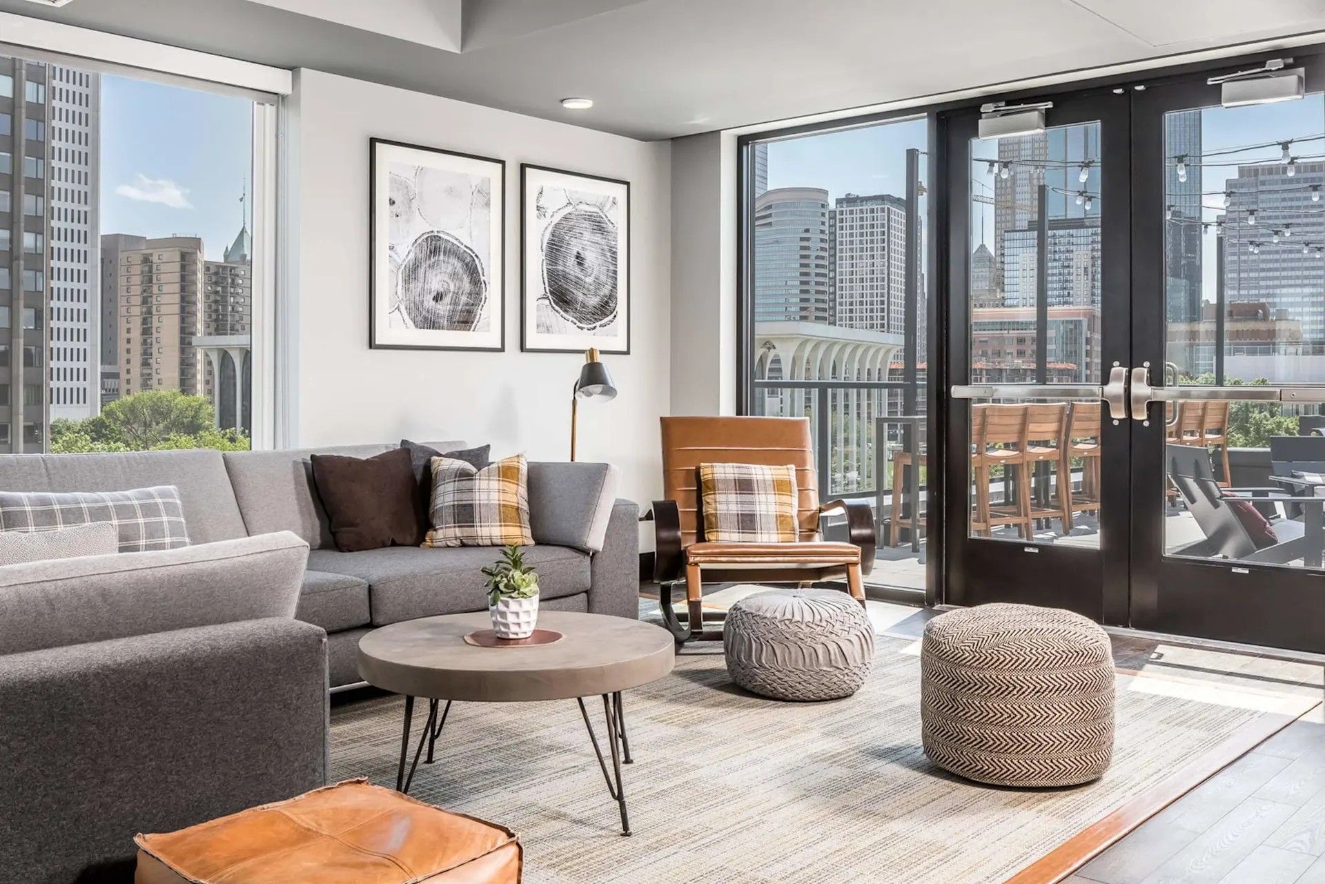 Modern apartment lounge with gray sofa, armchairs, two circular ottomans, coffee table, and patio view of city buildings.