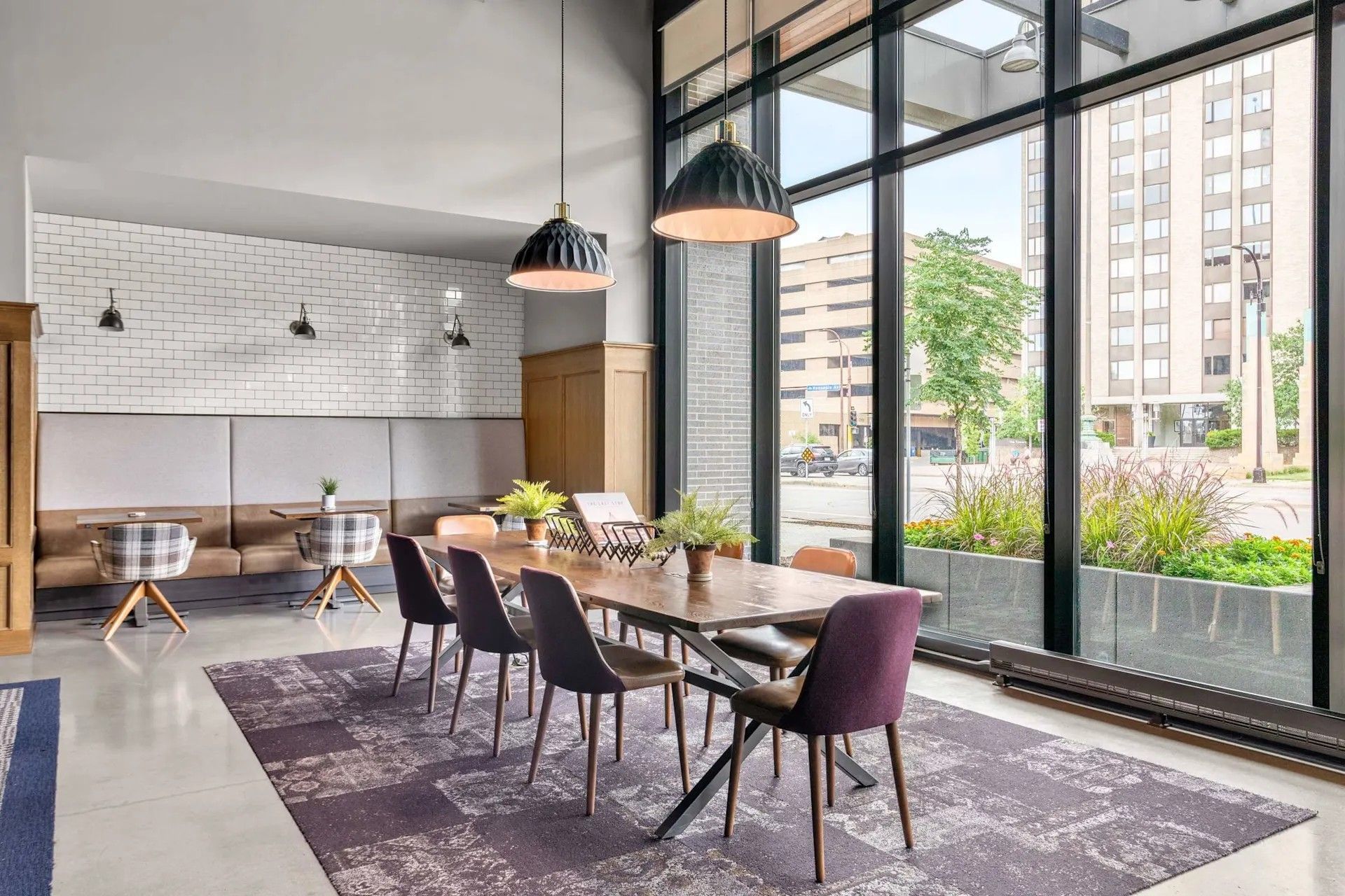 Modern lounge area with a long wooden table, purple and black chairs, and large floor-to-ceiling windows overlooking a street.