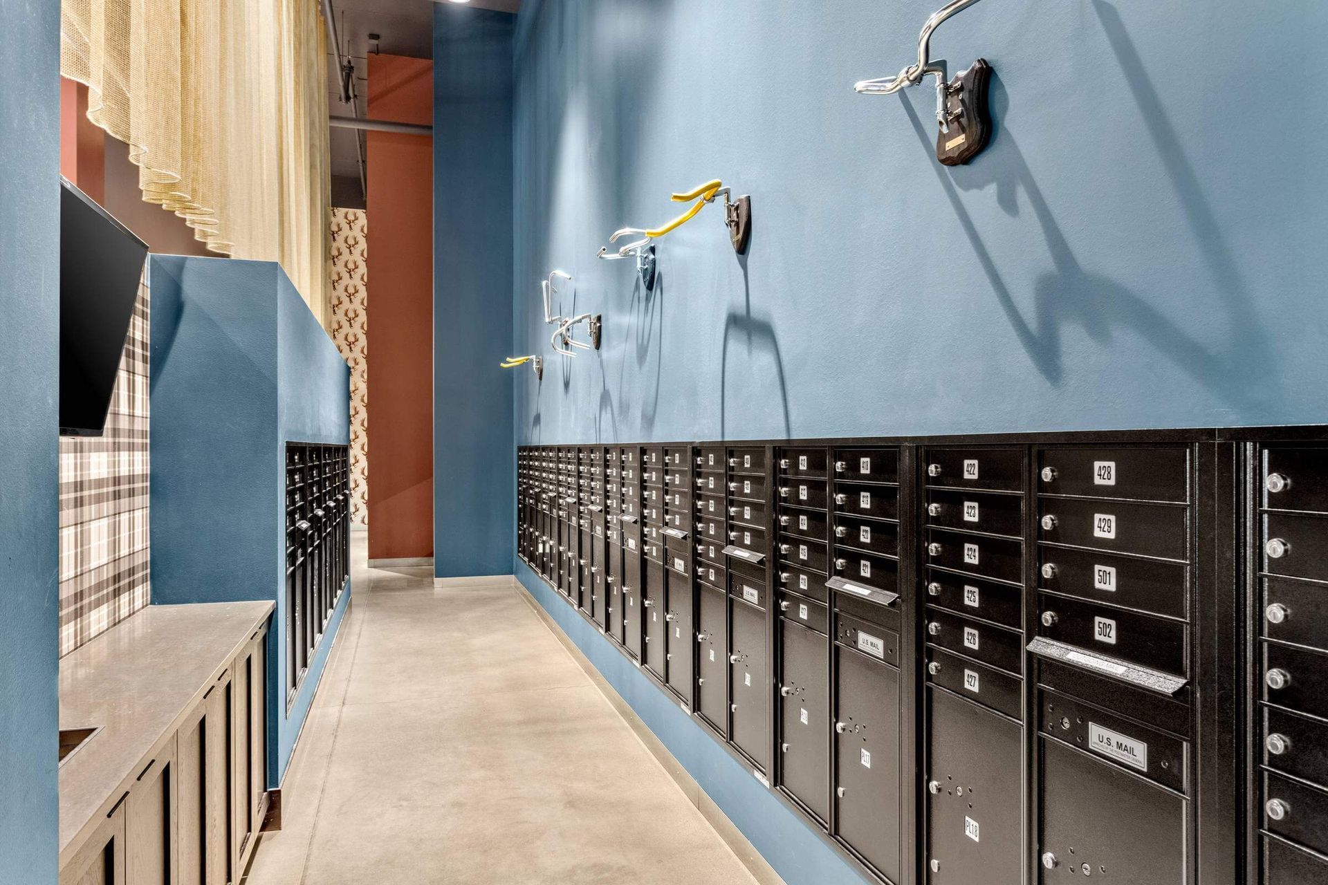 A long, narrow mailroom with blue walls, concrete floors, and rows of black mailboxes mounted on the right wall.