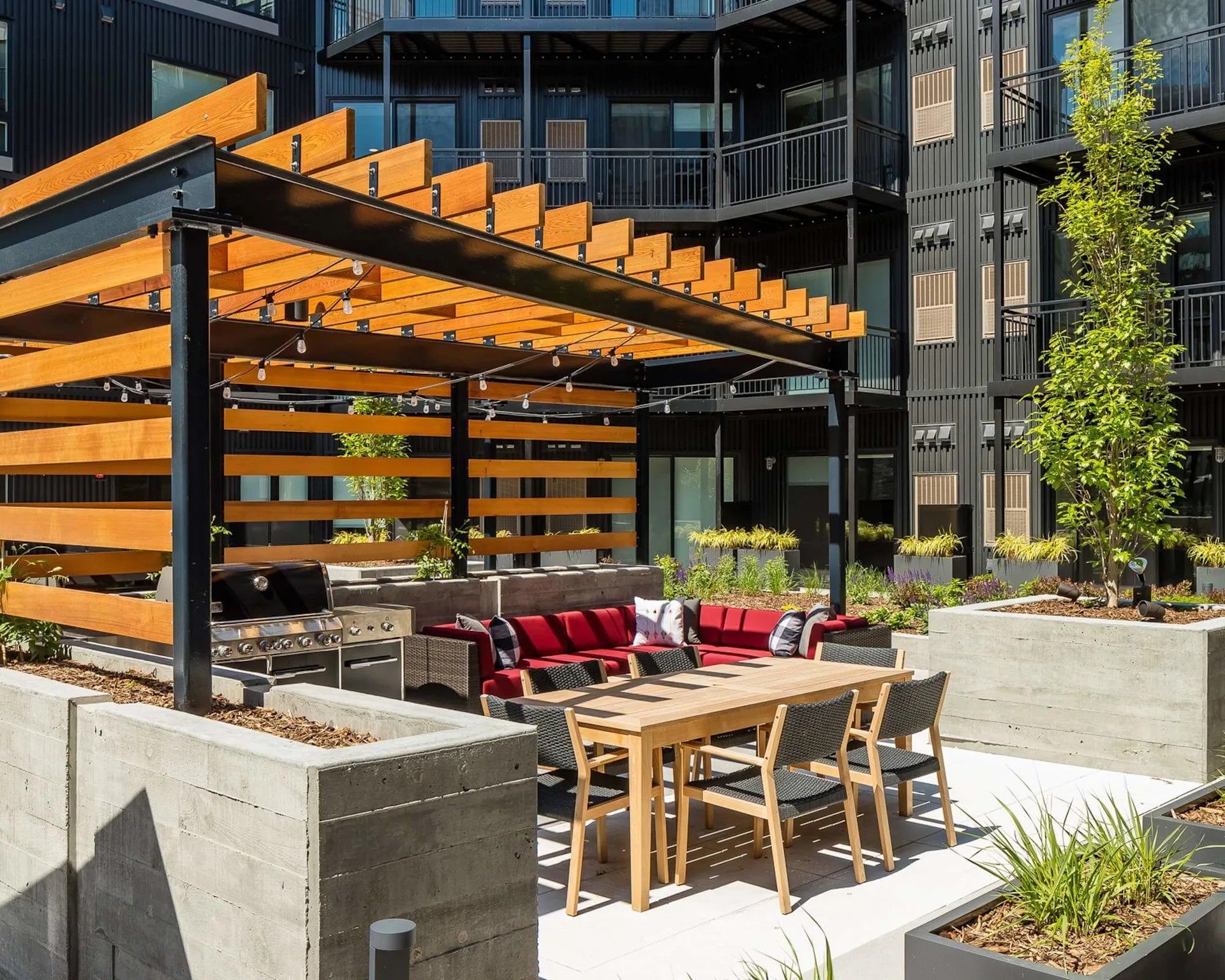 Outdoor patio with a wooden pergola over a dining table, chairs, and red cushioned seating against a modern dark building.