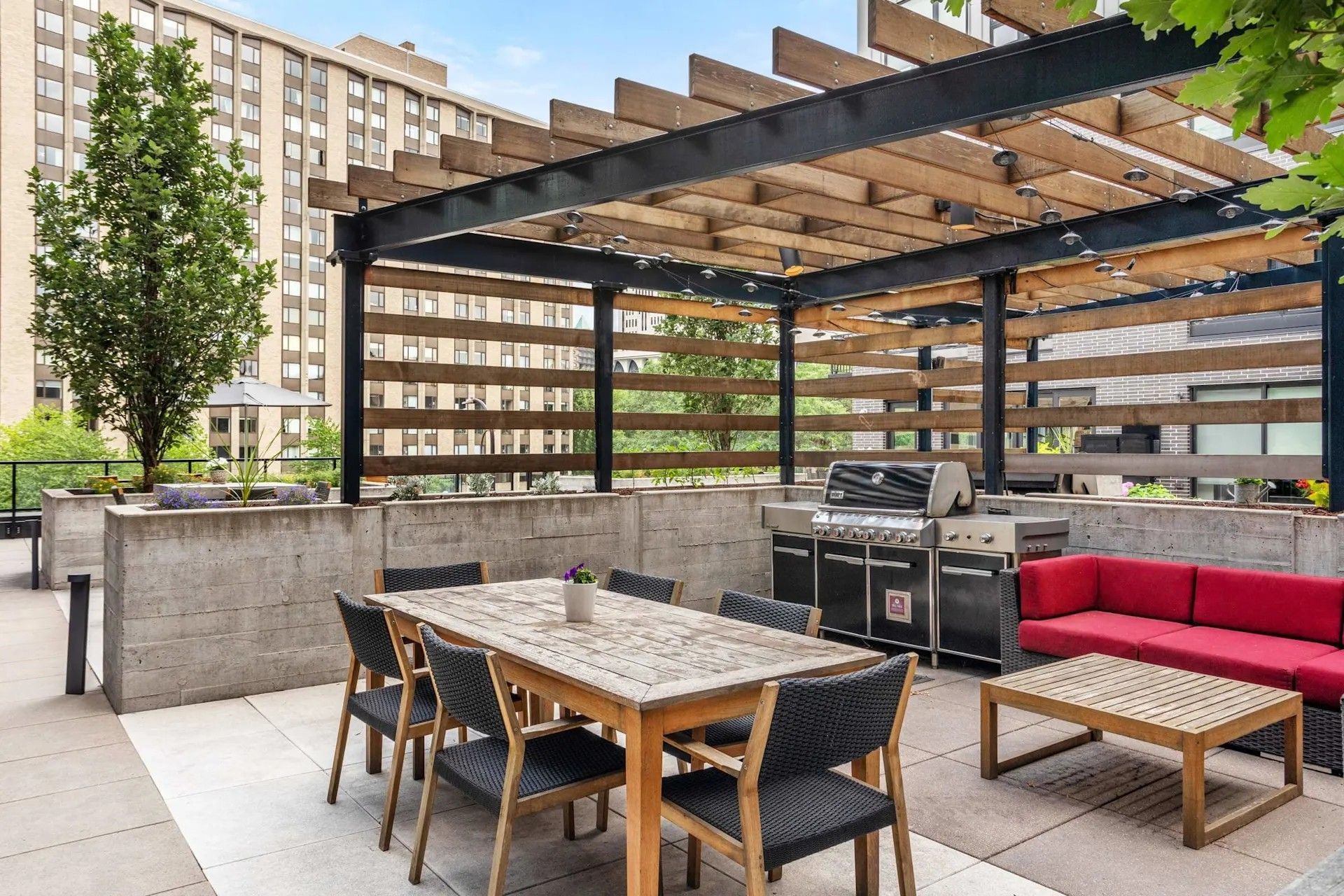 A rooftop patio featuring a wooden pergola, outdoor dining set, red sofa, and grill against a concrete wall.