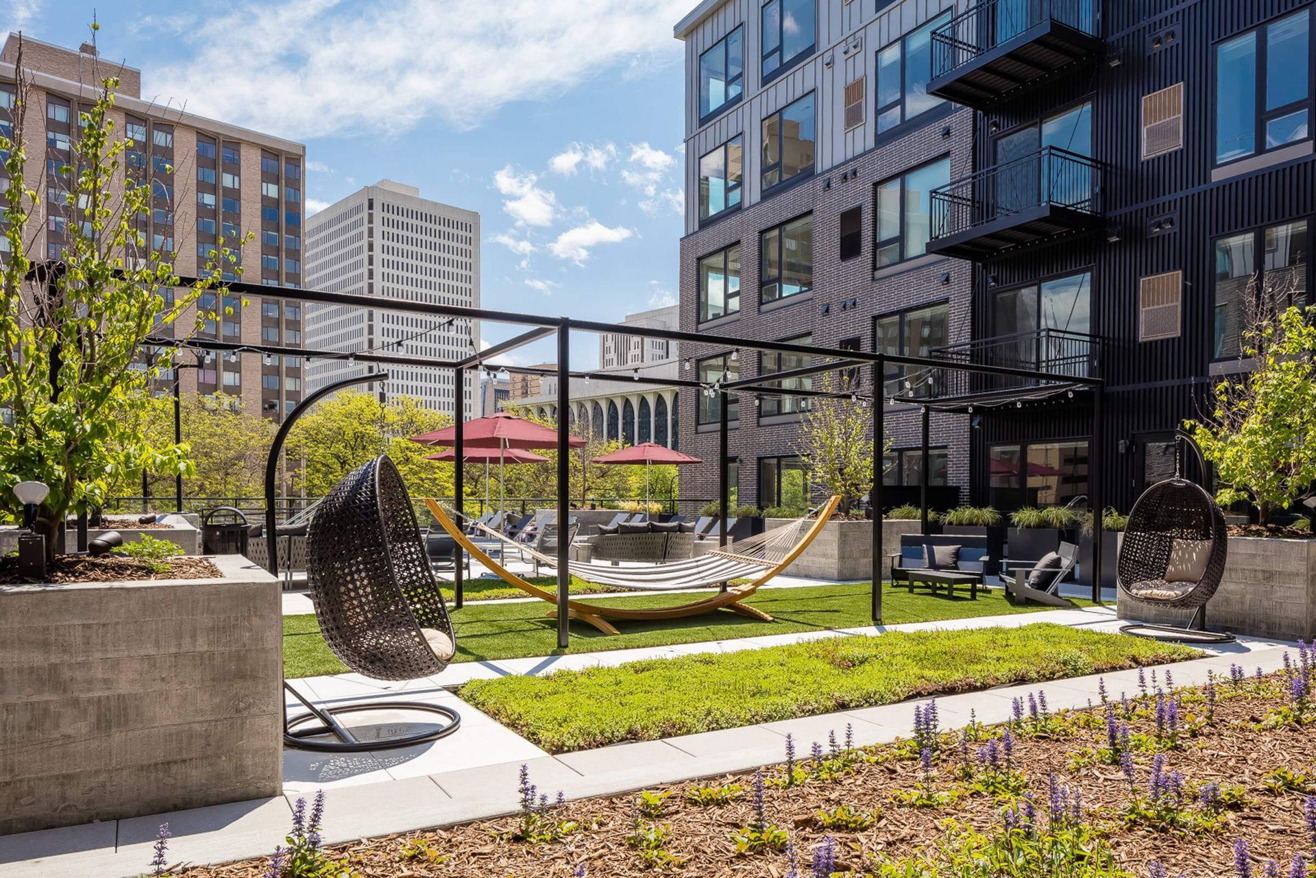 A sunny rooftop patio featuring a green lawn, a yellow hammock, hanging chairs, and planters against city buildings.