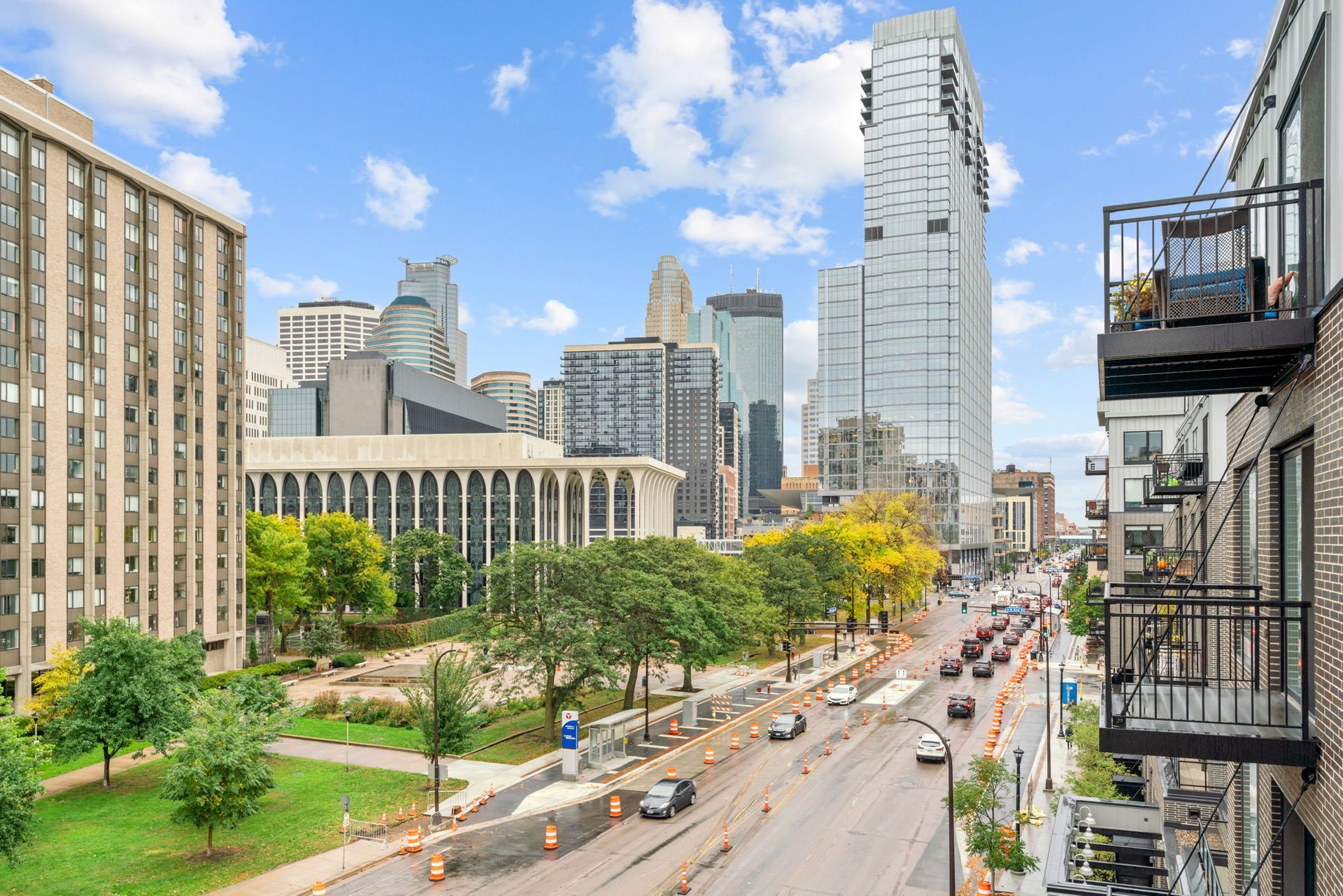 An elevated street-level view of a city center with skyscrapers, tree-filled parks, and traffic on a sunny day.