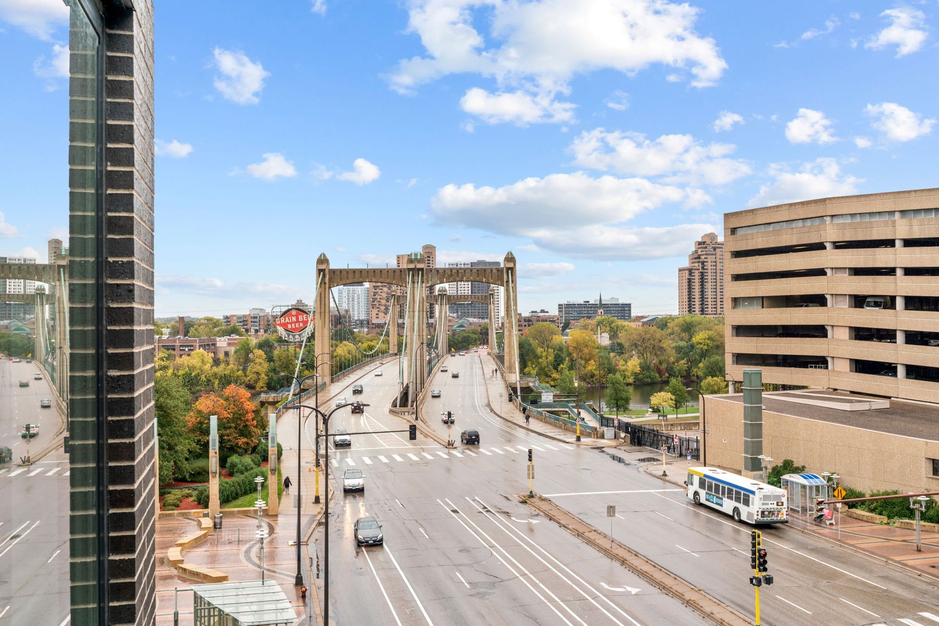 A bridge spans a wide city road on a cloudy day, with a bus and traffic below, surrounded by trees and a parking garage.