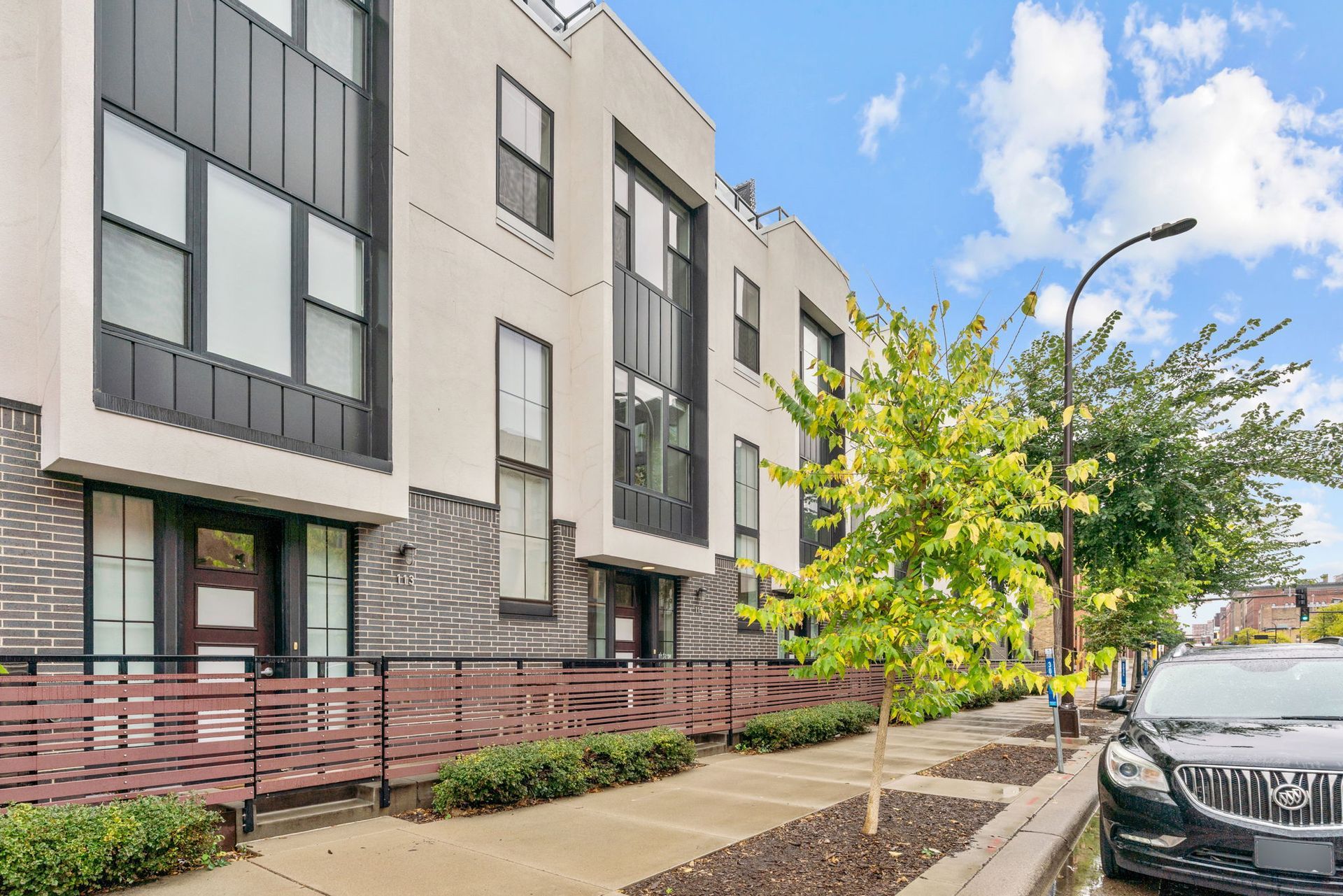 A modern multi-story townhome with a dark metal and light-colored facade, fenced front yard, and a car parked on the street.