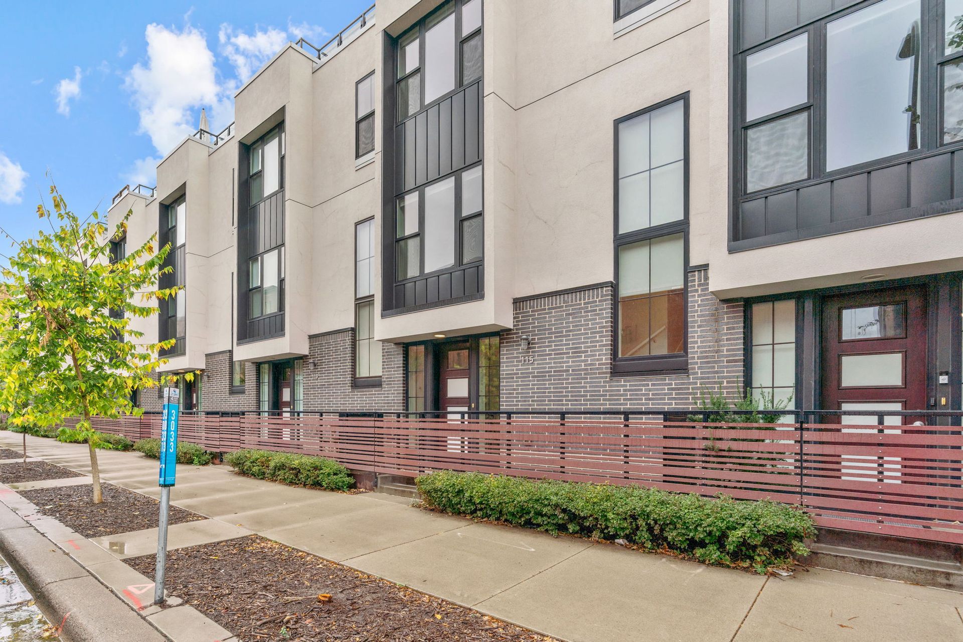 A row of modern, light-colored townhouses with metal trim, a red horizontal slat fence, and a sidewalk in front.