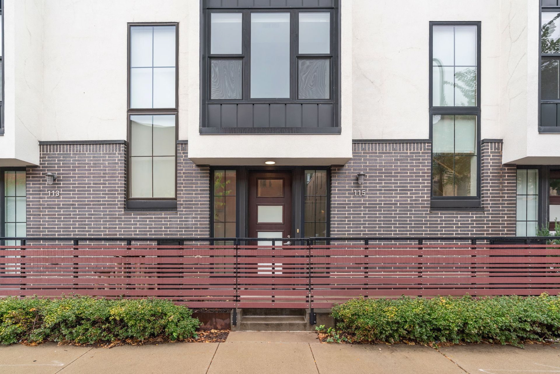 A modern townhouse entrance with white and dark brick exterior, large windows, a wooden front door, and a red metal fence.