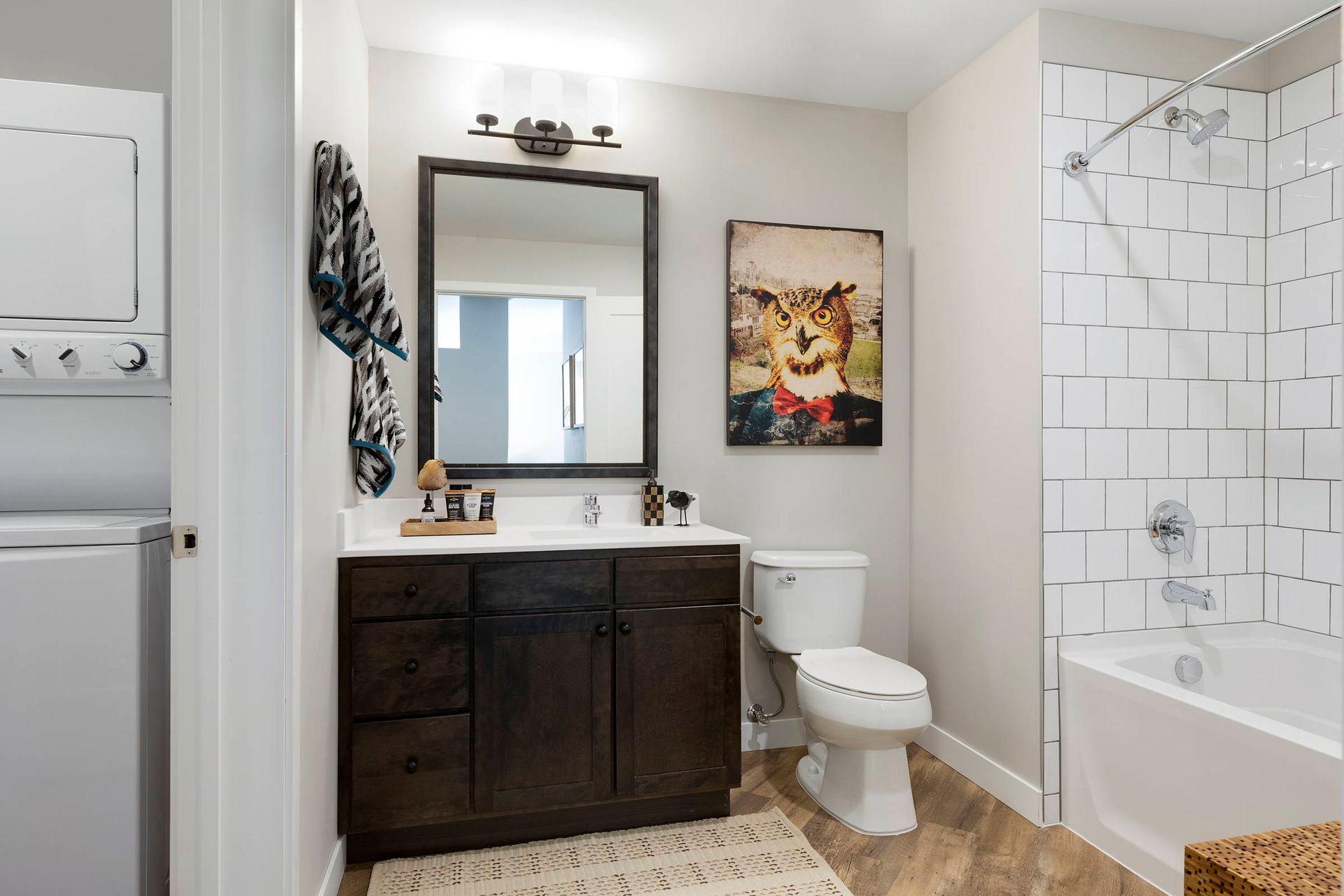 A bright bathroom here at Maverick with a dark wood vanity, a rectangular mirror, a toilet, a tub with white tile, and a stackable washer.