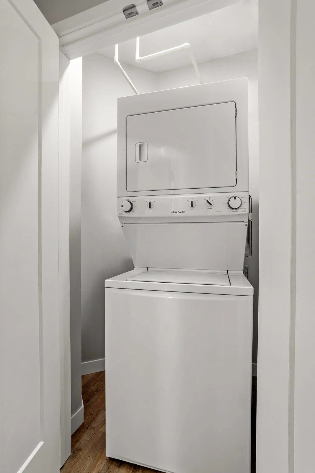 A white stacked washer and dryer unit located inside a narrow indoor laundry closet with wood flooring.
