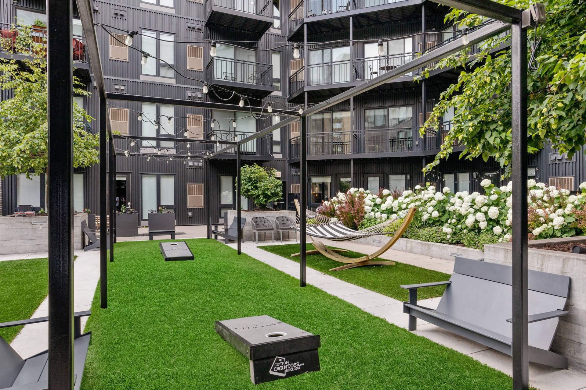 A courtyard with a lawn , benches , chairs , and a cornhole board.