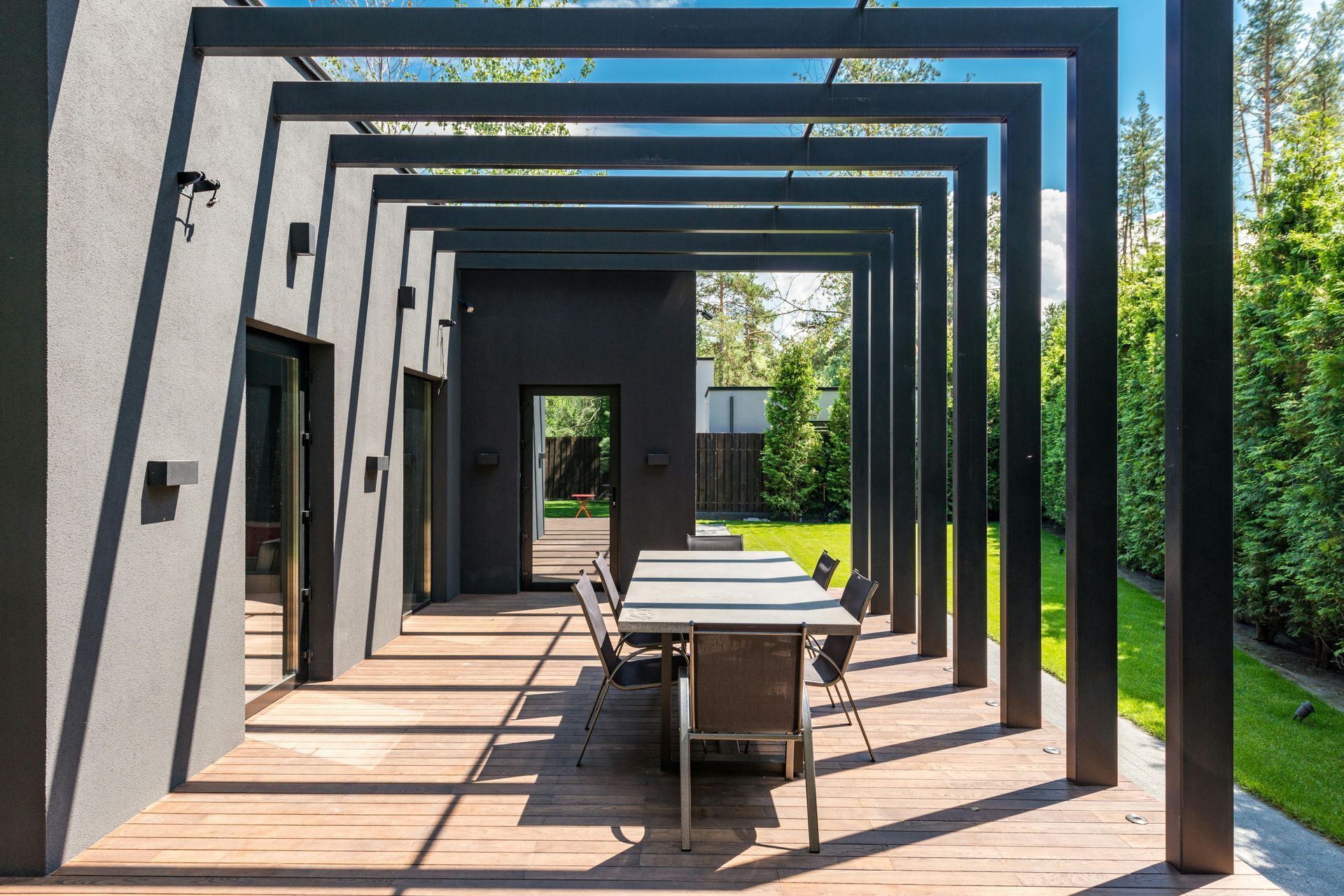 Modern outdoor dining area with a long table under a dark-framed pergola, next to a black exterior wall, green lawn in the background.