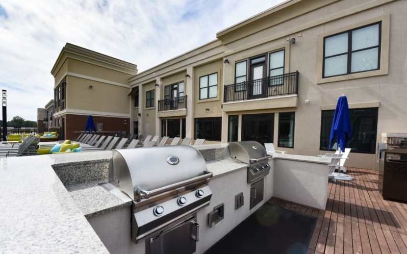 Outdoor grilling station near a pool at an apartment complex. Stainless steel grills, blue umbrellas, and beige building.