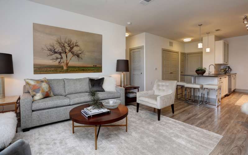 Living room with gray sofa, white chair, and art; open to kitchen with bar stools.
