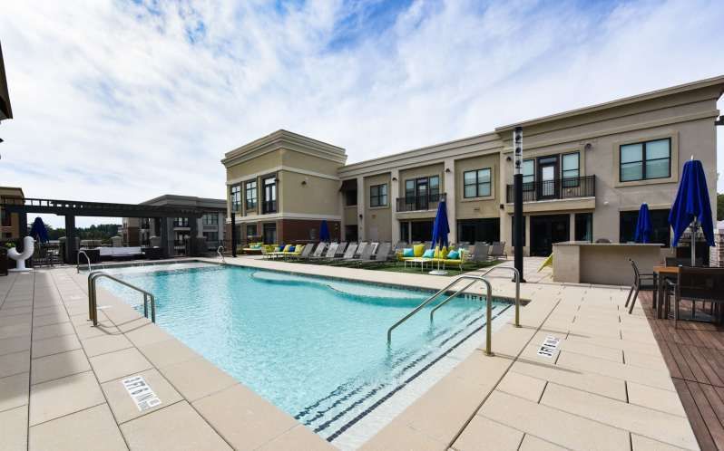 Swimming pool and patio at an apartment building, with lounge chairs, umbrellas, and clear sky.