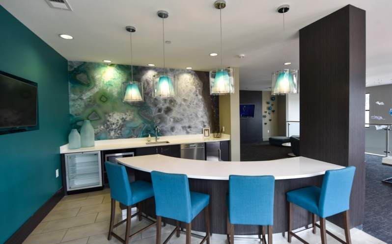 A modern bar area with teal accents, a white countertop, blue stools, and pendant lights.