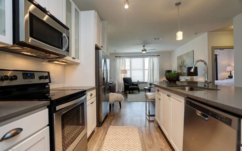 Modern kitchen with stainless steel appliances, white cabinets, and a view into a living room with natural light.