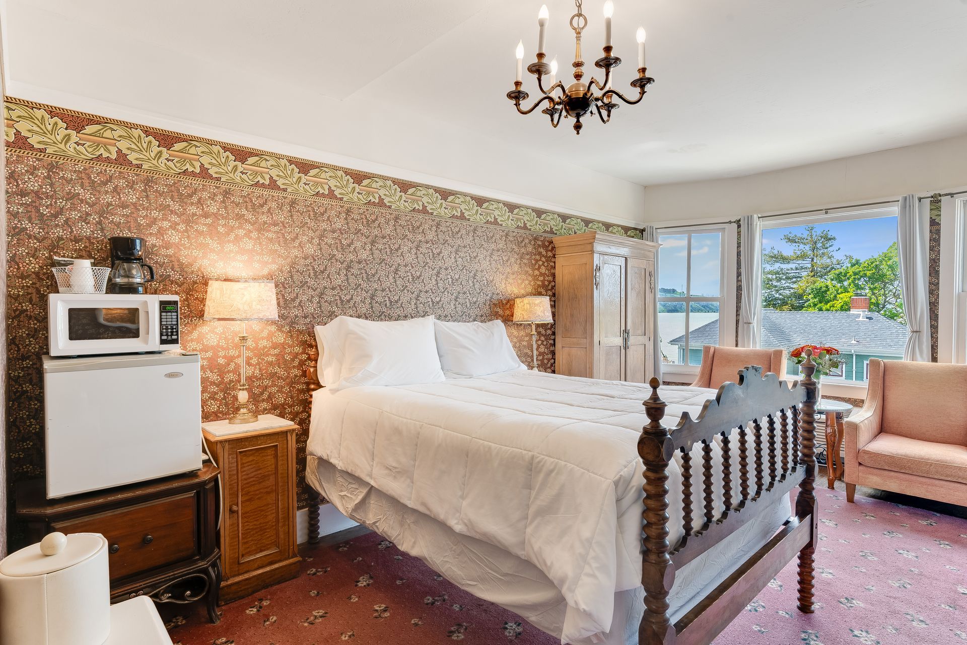 Bedroom with a bed, small fridge, microwave, and pink chairs. Brown wallpaper, and ornate chandelier.
