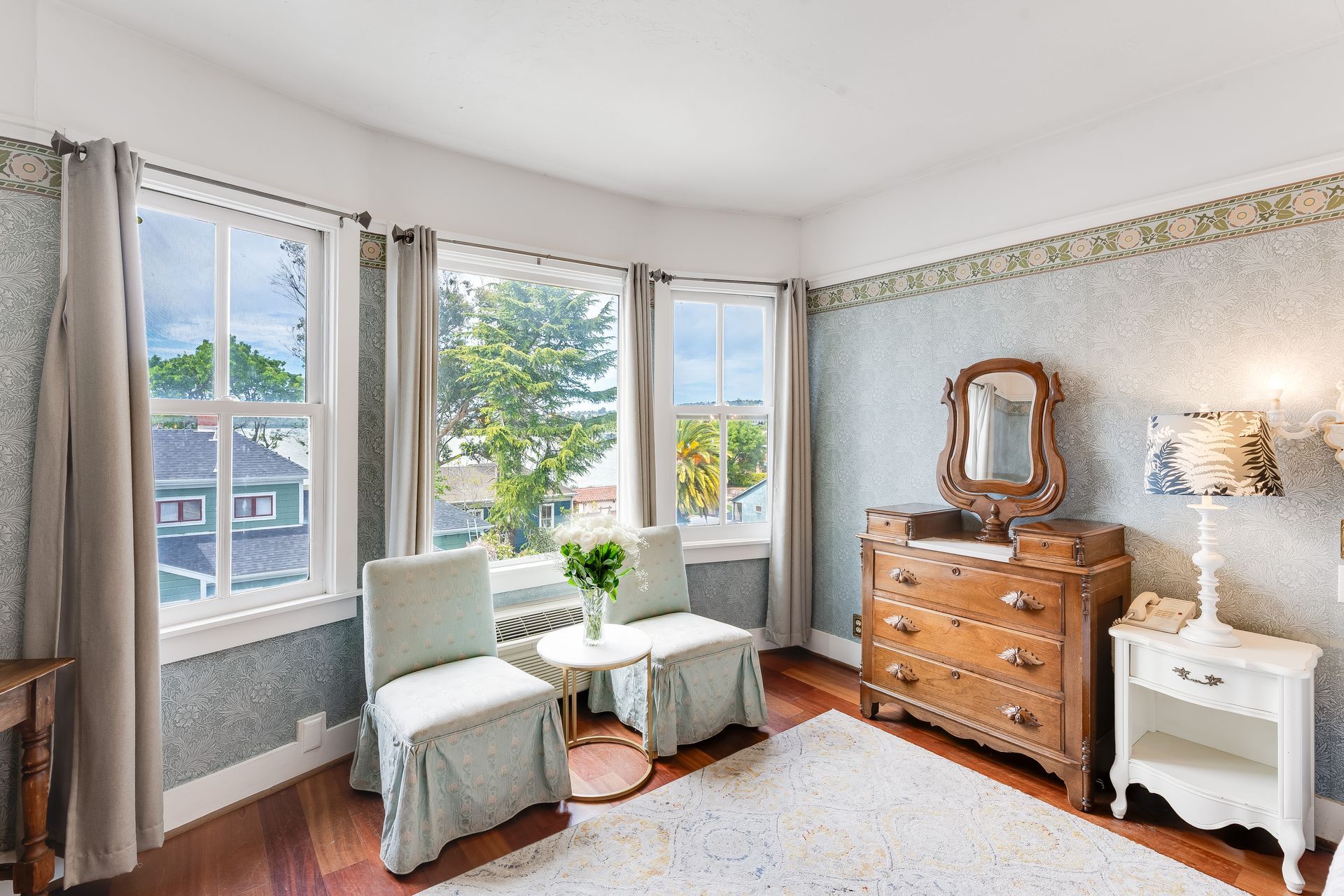 Bedroom with bay windows, antique furniture, floral wallpaper, and two chairs.