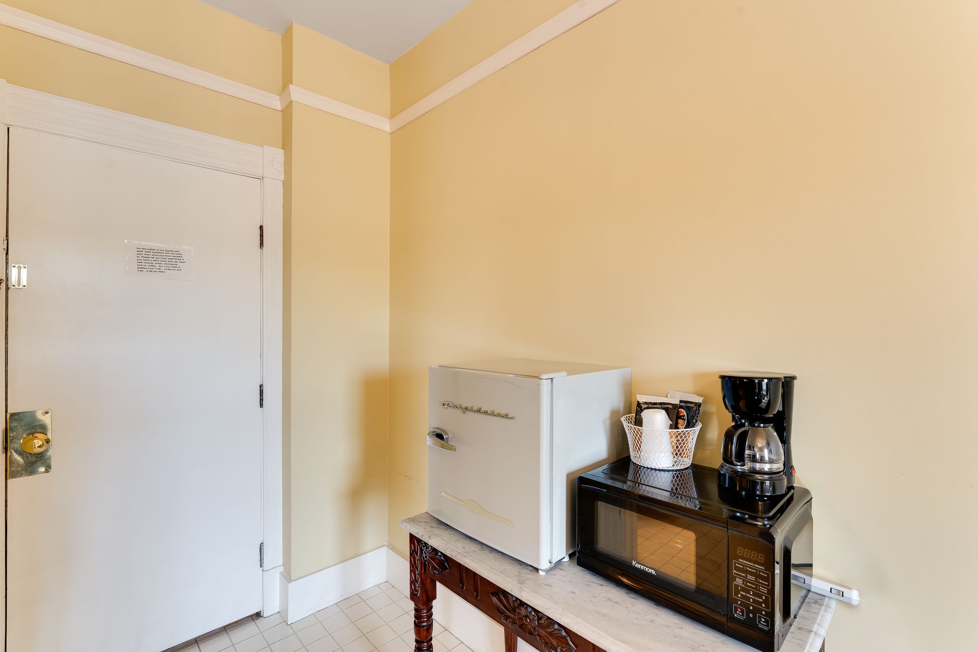 A small room corner with a white mini-fridge, microwave, coffee maker, and door. Yellow walls, wooden table.