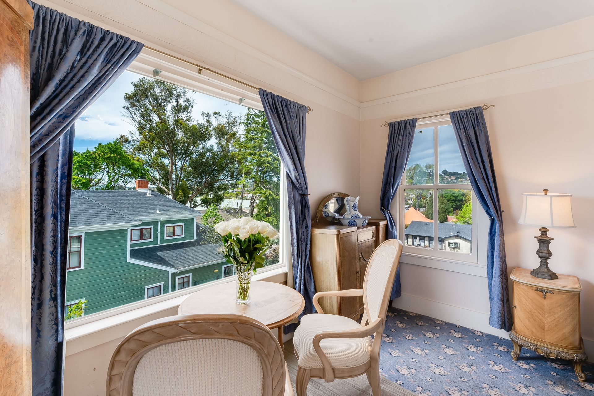 A light-filled room with a view of a green house. Blue curtains, white furniture, a table with flowers.