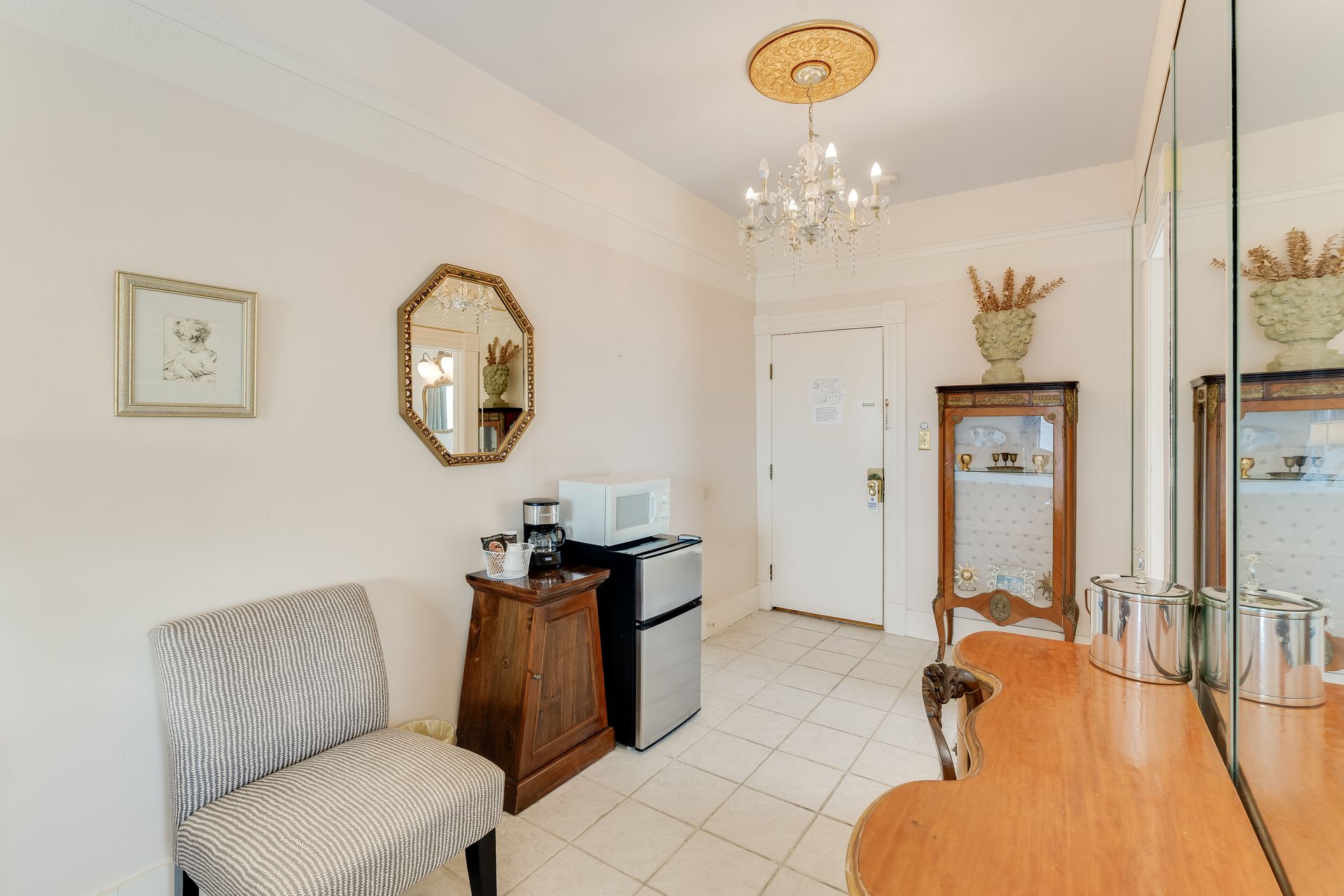 A hotel room with a chair, table, mirrors, microwave, coffee maker, and a door.